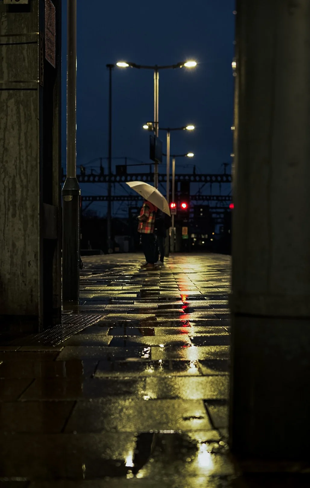 Two people stand on a wet city sidewalk at night, one holding an umbrella, illuminated by streetlights and distant red train signals.