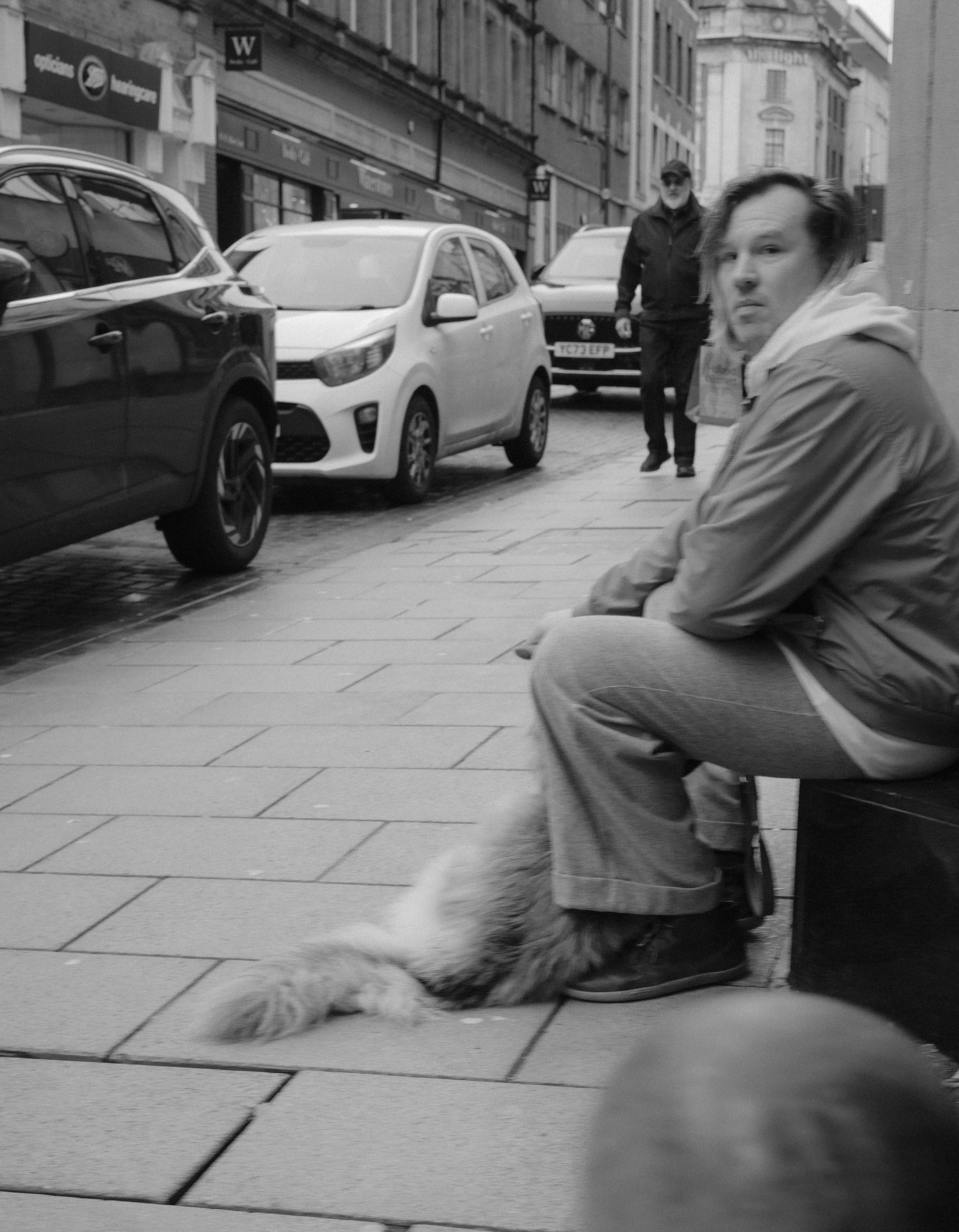 A black and white photo of a city sidewalk with parked cars, a man sitting on a bench, and a woman with a dog partially visible at the bottom of the frame.