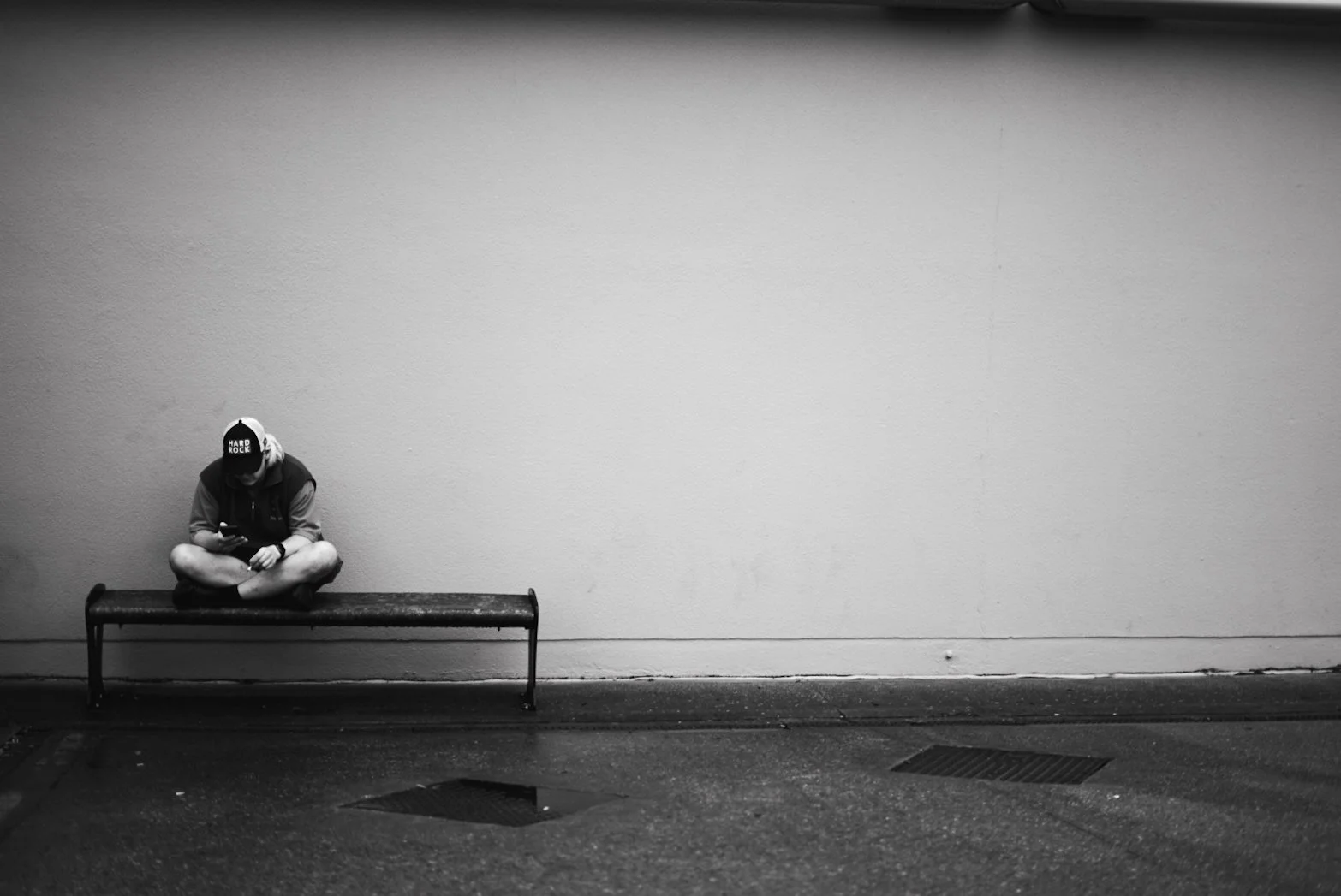 A person sitting cross-legged on a bench in front of a plain wall, wearing a hoodie, shorts, and a cap, looking at their phone in black and white.