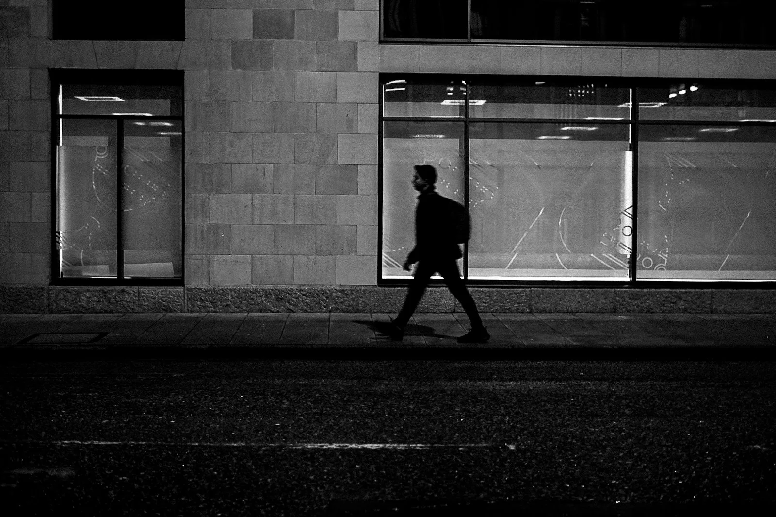 A silhouette of a person walking past a large window on a city street at night, illuminated by streetlights, with reflections visible in the window.