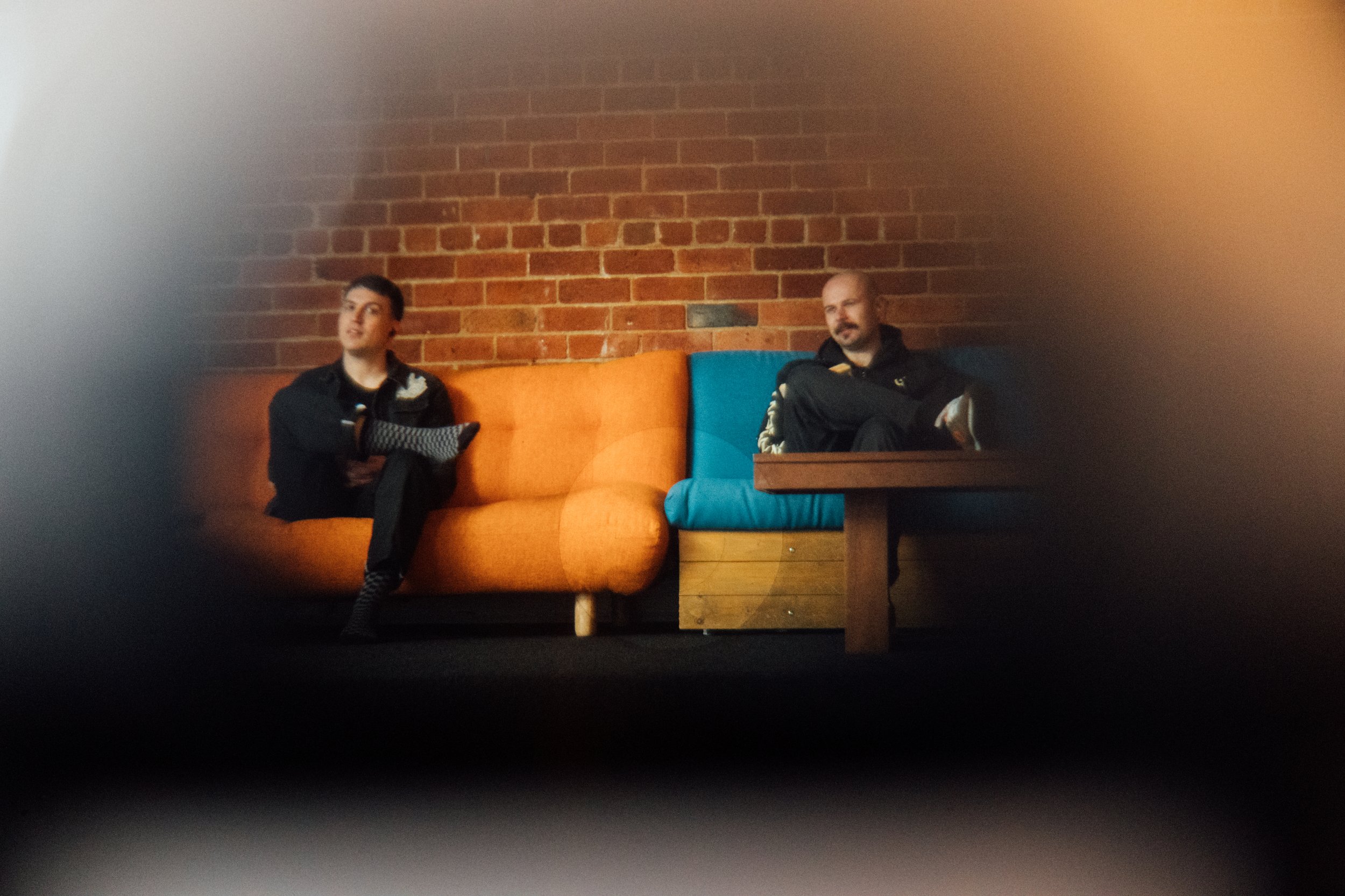 Two men sitting on colorful couches, one orange and one blue, against a brick wall in a casual indoor setting.
