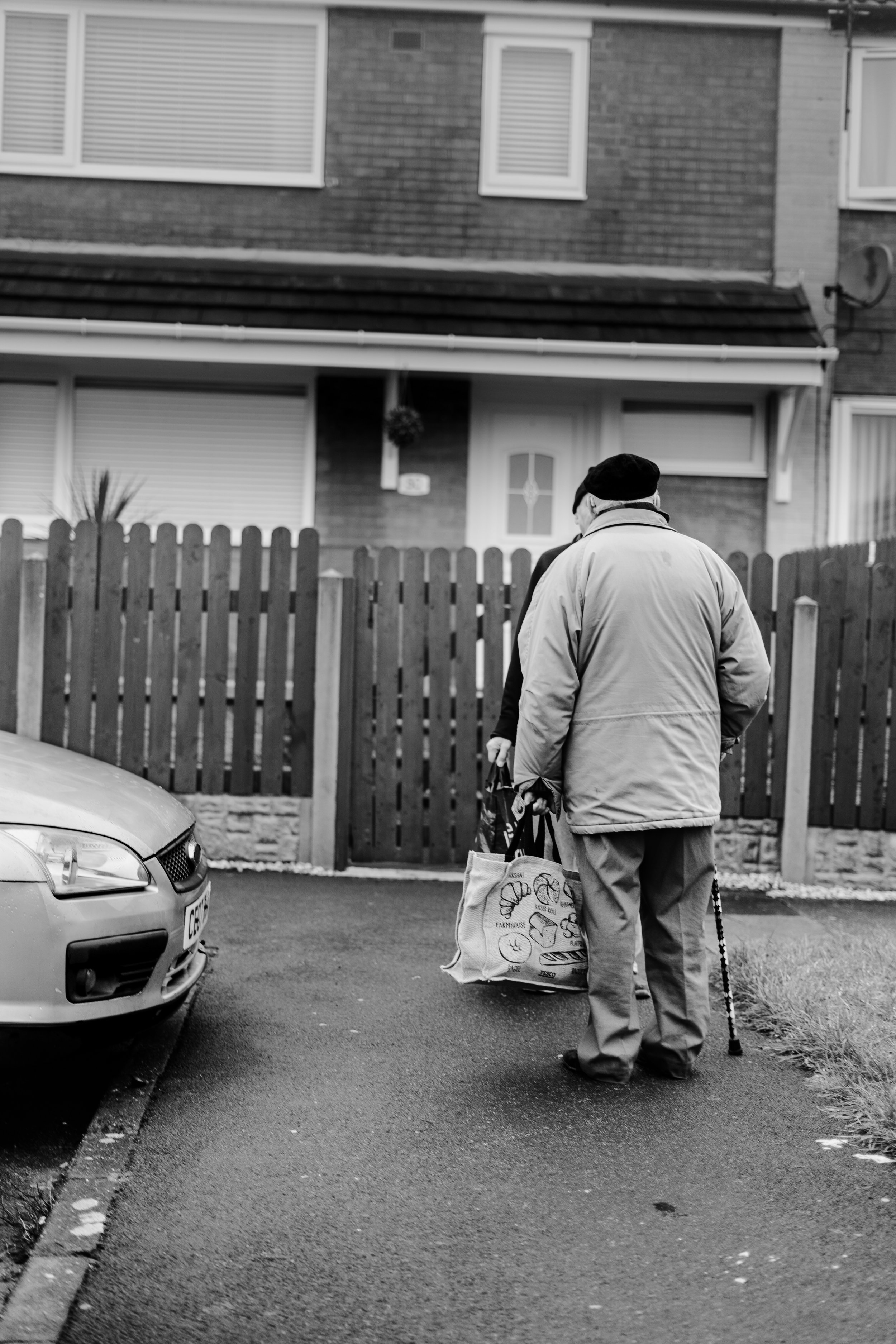 An elderly man with a cane is walking on a sidewalk, carrying grocery bags, with a parked car on his left and a residential house with a wooden fence in the background.