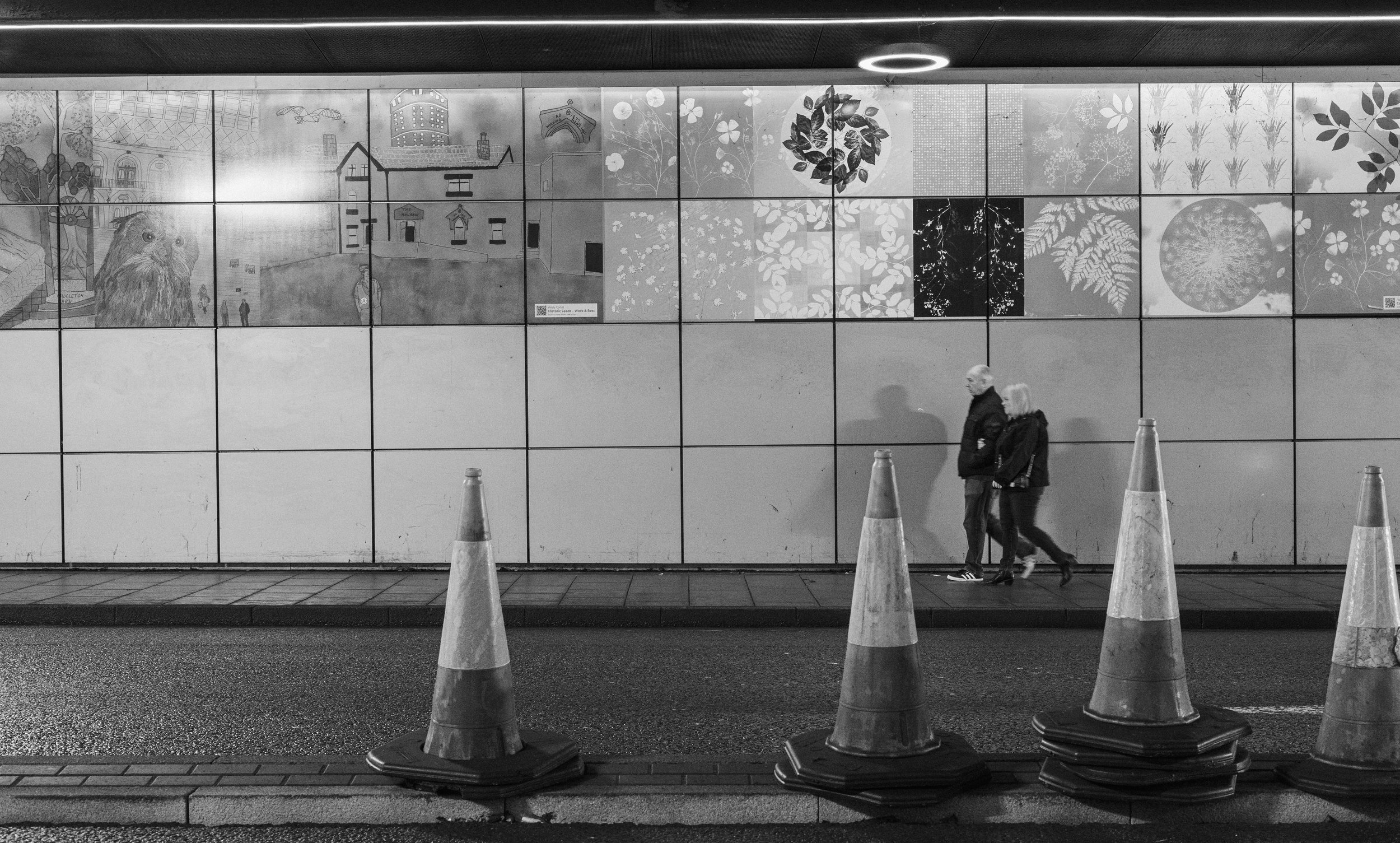Black and white photo of two people walking past a large wall mural under a city overpass, with traffic cones in the foreground.