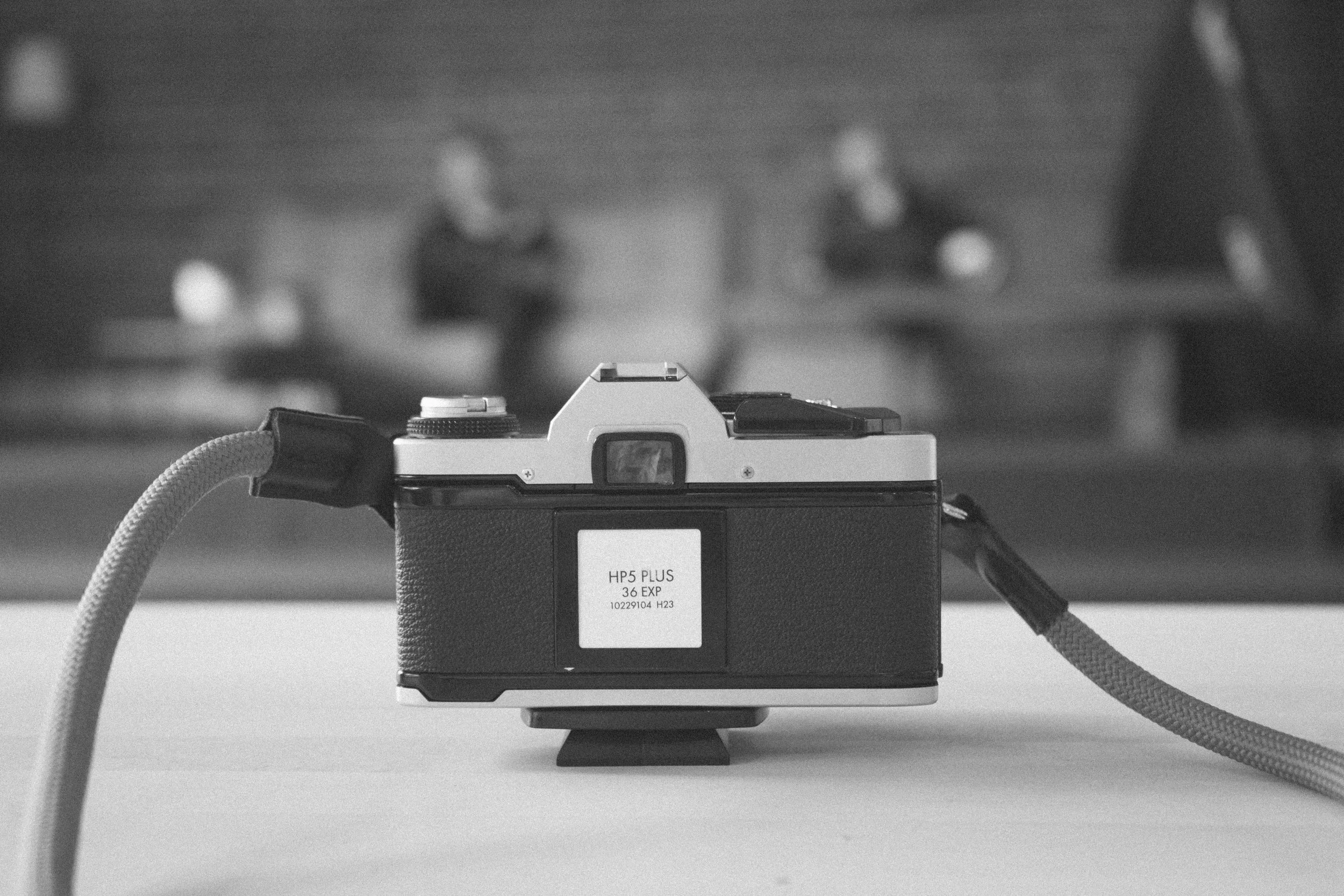 A vintage camera positioned on a table with two cables attached, one on each side. Two blurred individuals are visible in the background, sitting on a couch in a room with a brick wall.