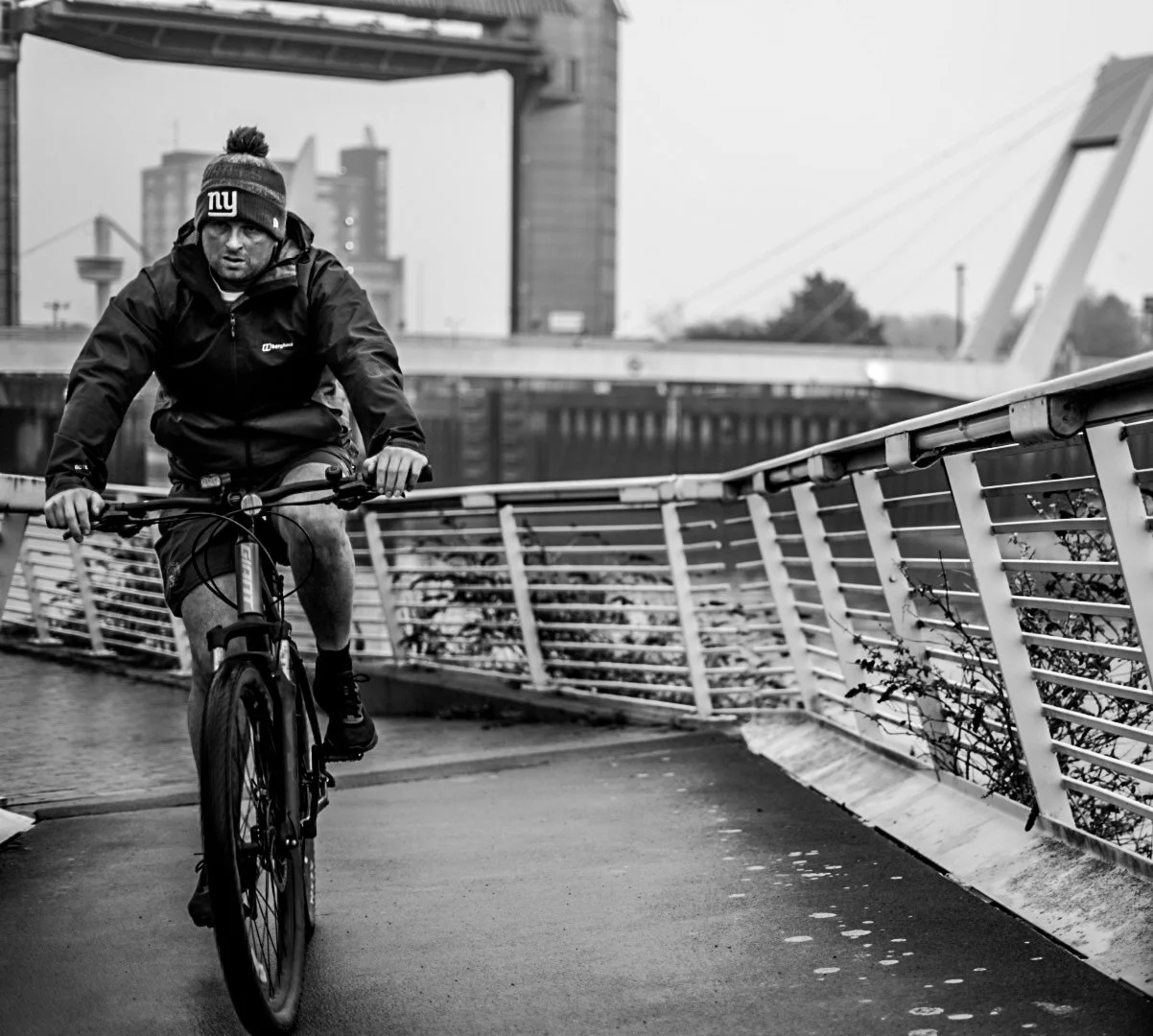 A man biking on a city bridge with buildings in the background, dressed in a black jacket, shorts, and a beanie with a sports logo.
