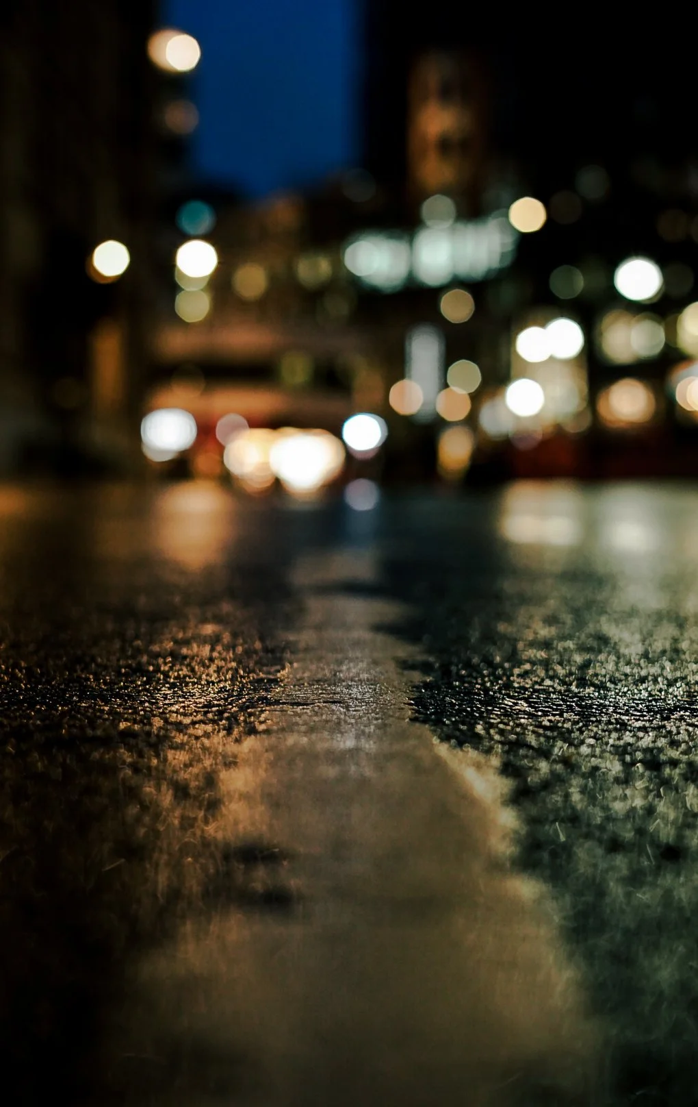 Blurred city street at night with city lights and cars in the background, viewed from a low angle on a wet road.