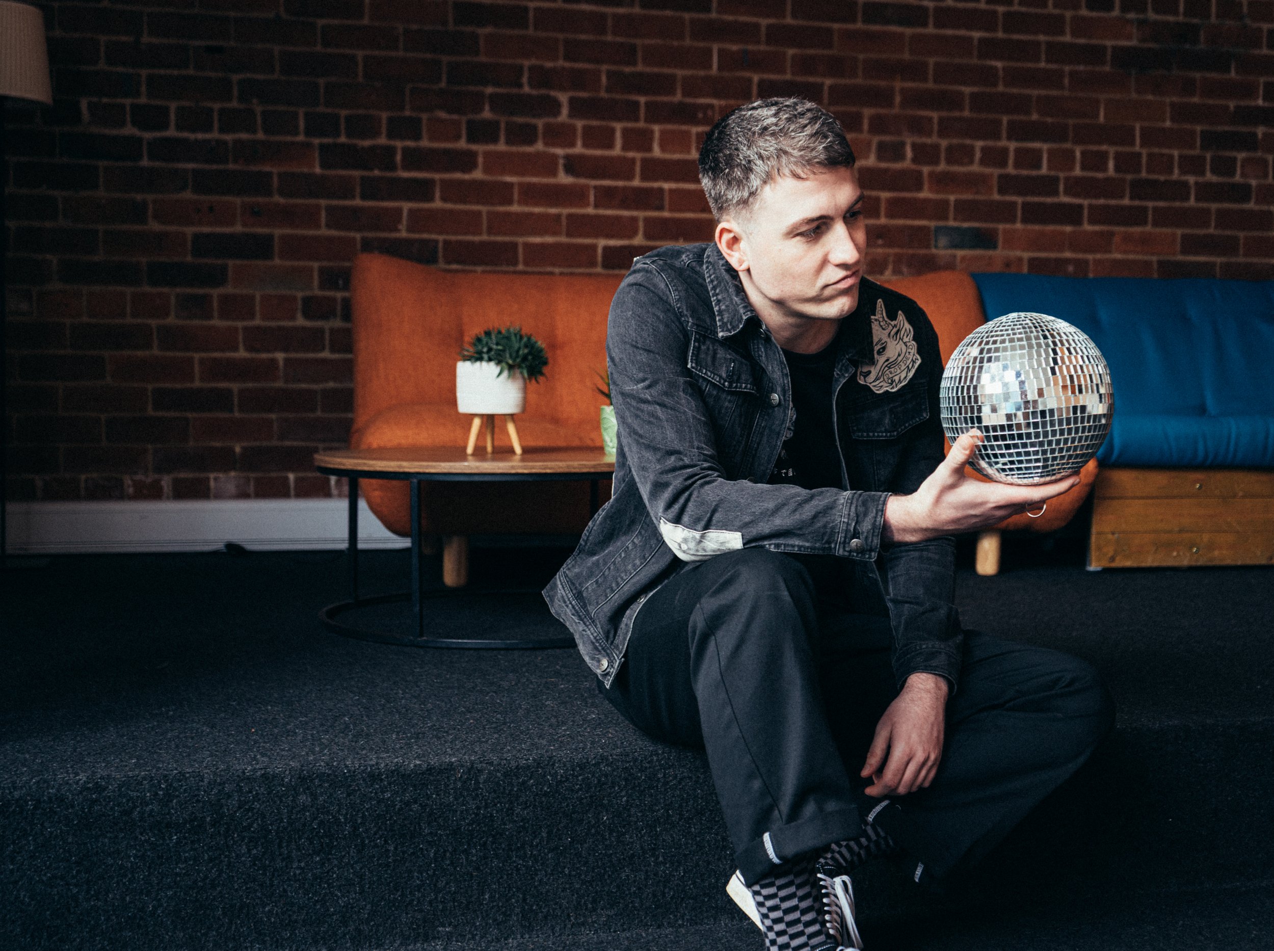 A young man with short hair, wearing a black denim jacket and black pants, sits on a dark carpeted floor in a room with a brick wall background. He is holding a small disco ball in his right hand and looks at it with a thoughtful expression. Behind h