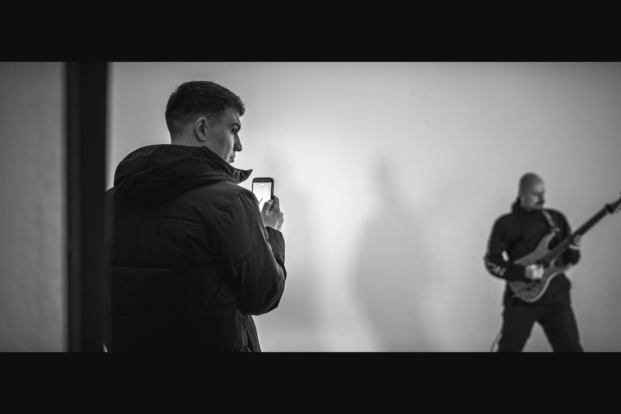 A young man in a black jacket taking a photo or video with his smartphone, while another person playing an electric guitar is in the background. The scene is in black and white.