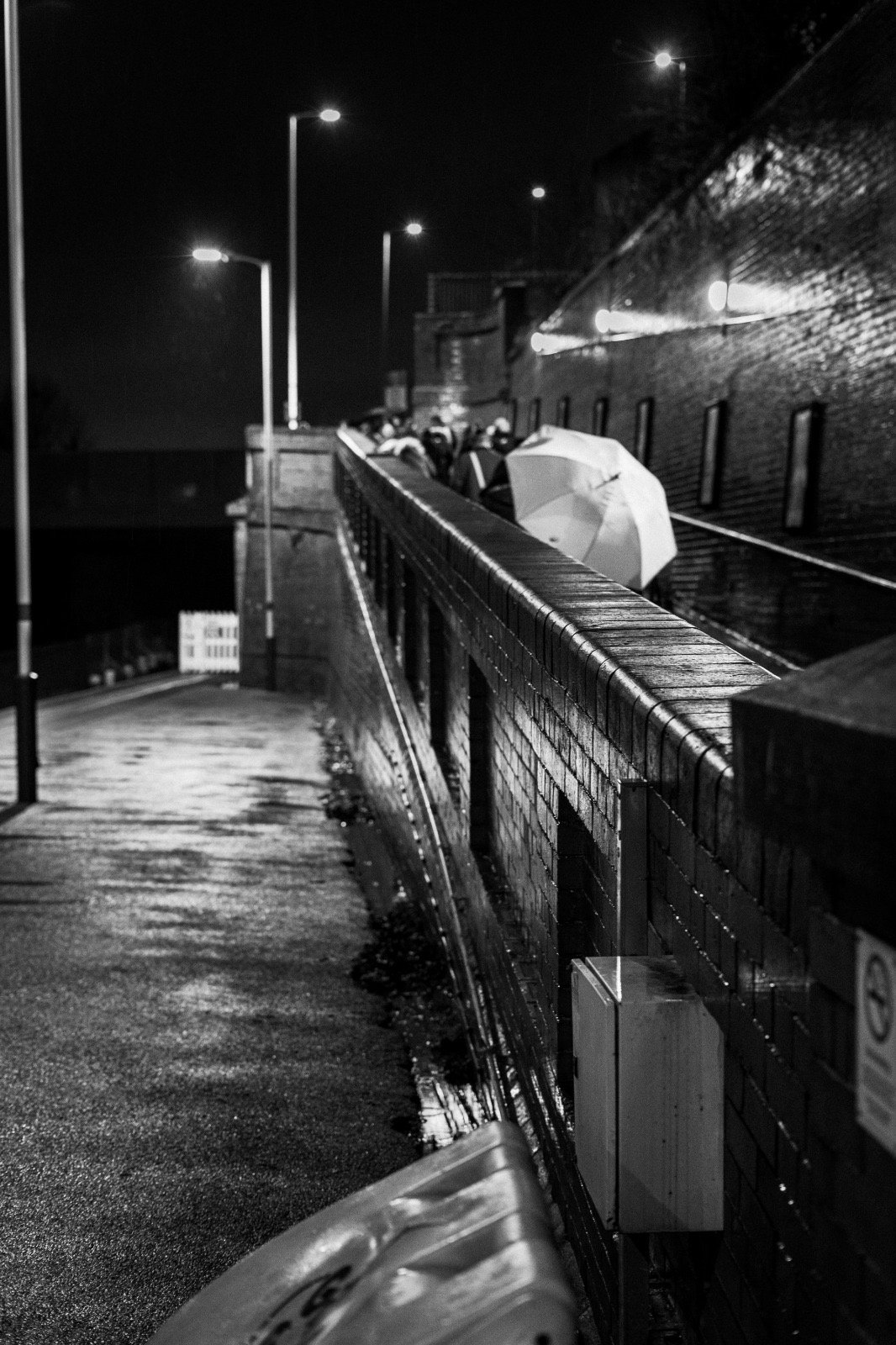 Nighttime scene with a wet, empty walkway illuminated by streetlights, a high brick wall on the right, and a group of people with umbrellas in the distance.