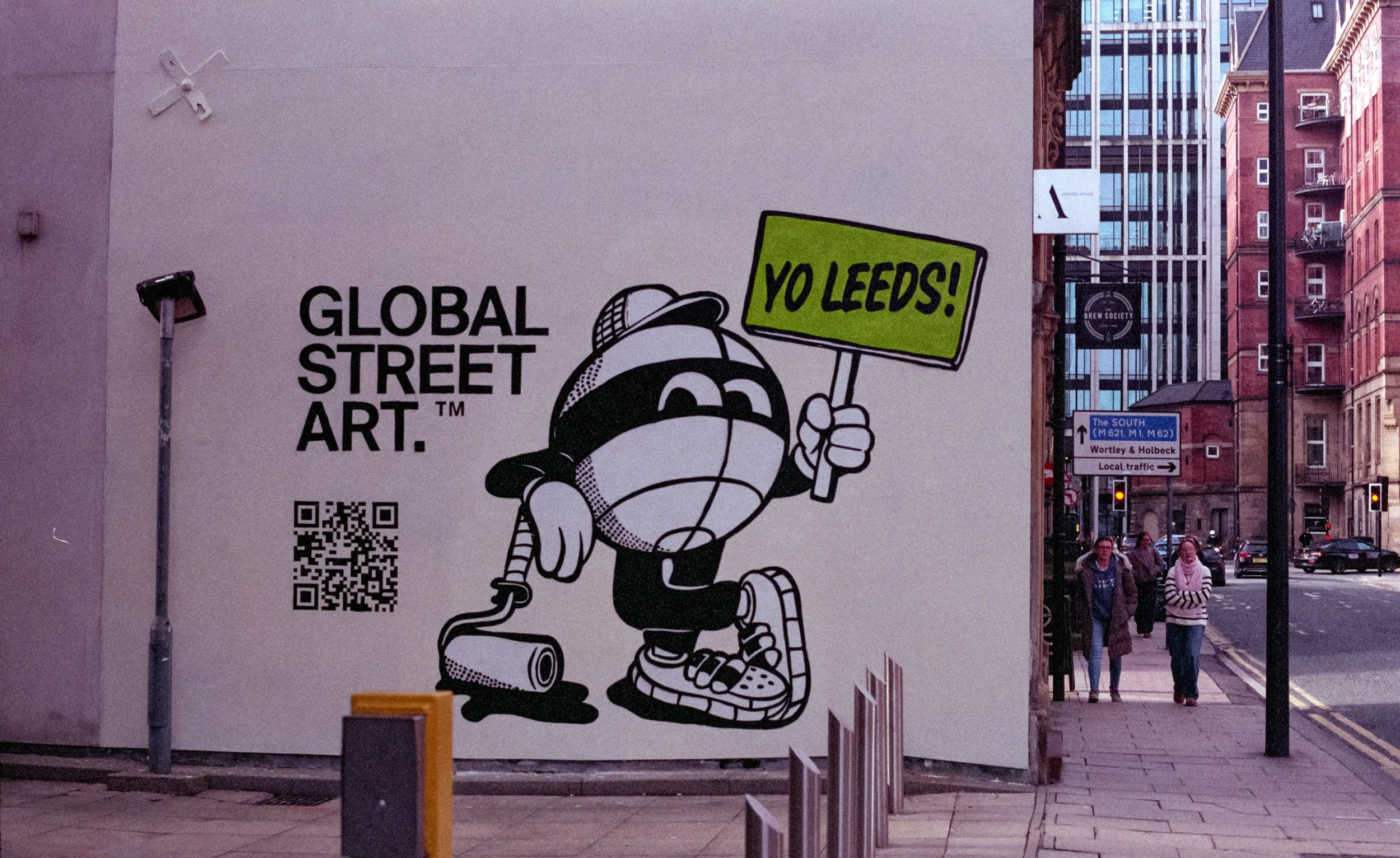 Street art poster with a cartoon character holding a sign that says "YO LEEDS!" and a speech bubble, on a city sidewalk with pedestrians and buildings in the background.