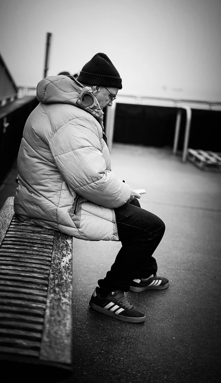 A man wearing a beanie, glasses, and a puffy jacket sitting on a wooden bench looking at his phone on an outdoor deck or patio.