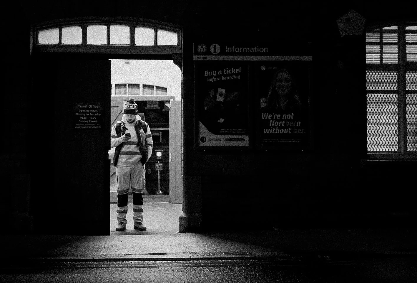 A person dressed in a striped sweater, shorts, and a beanie, standing in a train station entryway, looking at their phone.