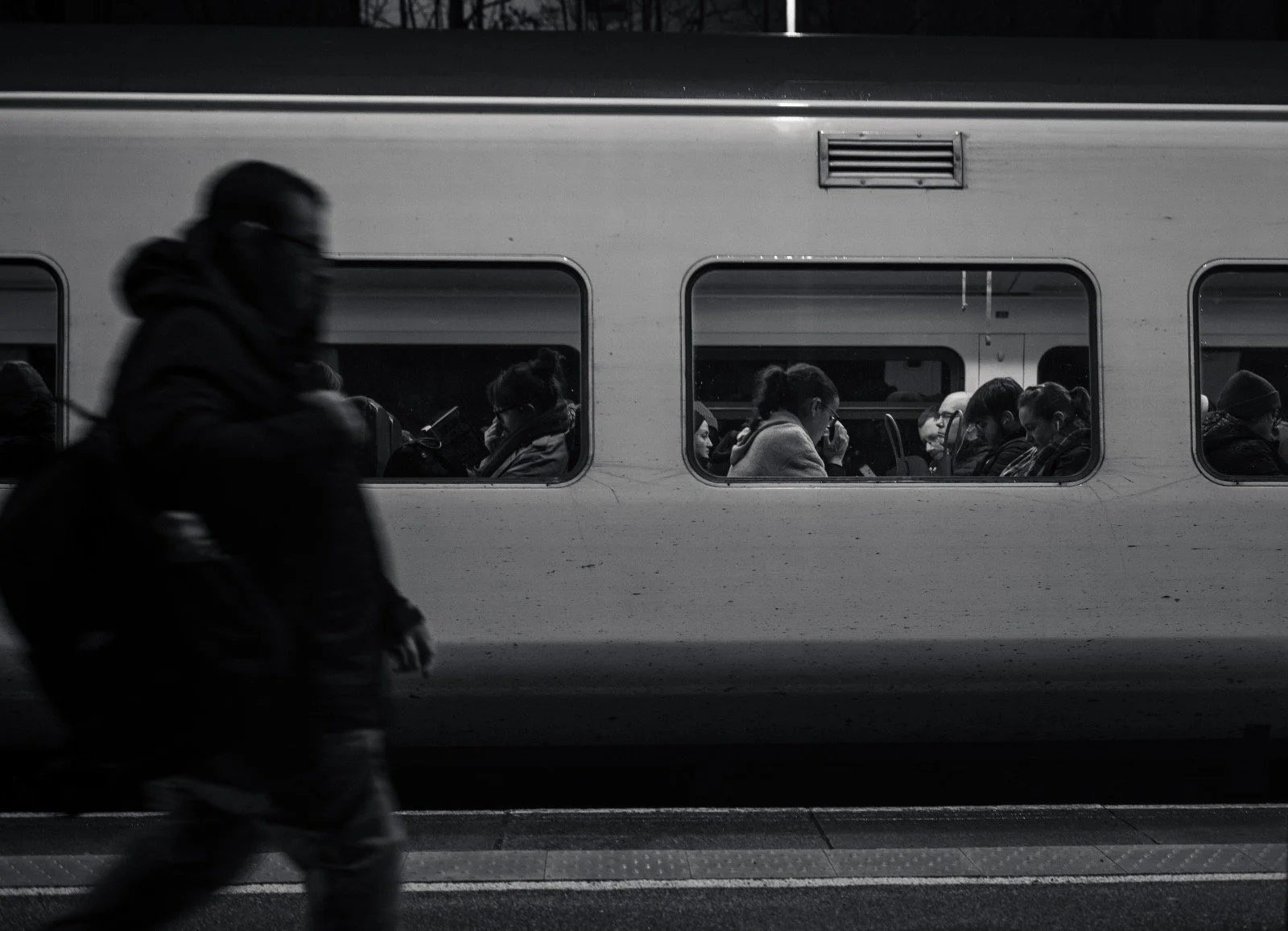 People sitting inside a train, viewed through the windows, with a blurred person walking past in the foreground. The image is in black and white.