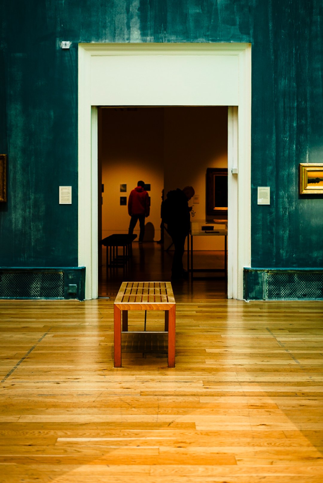 An art gallery with an empty wooden bench in the foreground, a large doorway leading into a dimly lit room with people viewing artwork, and framed paintings on the wall.
