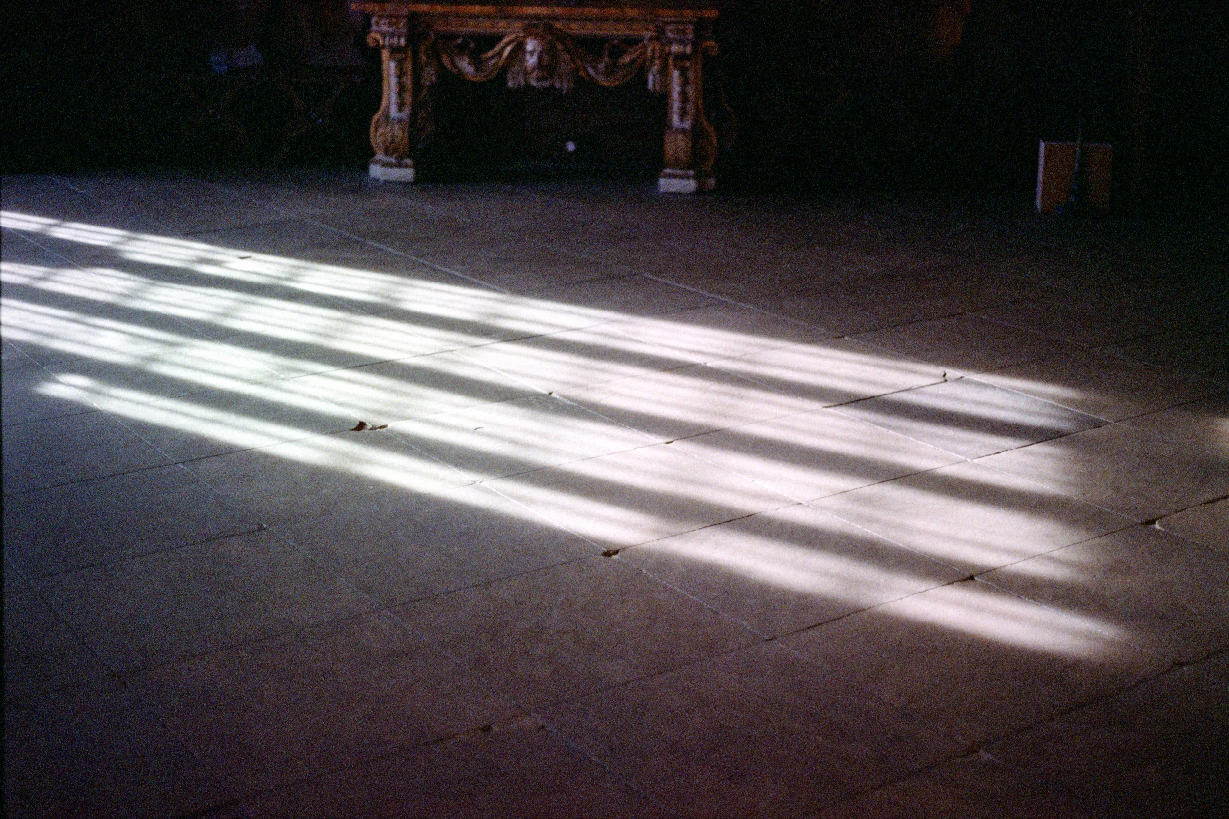 Sunlight streaming through window creating striped shadows on a tiled floor, with ornate furniture in the background.