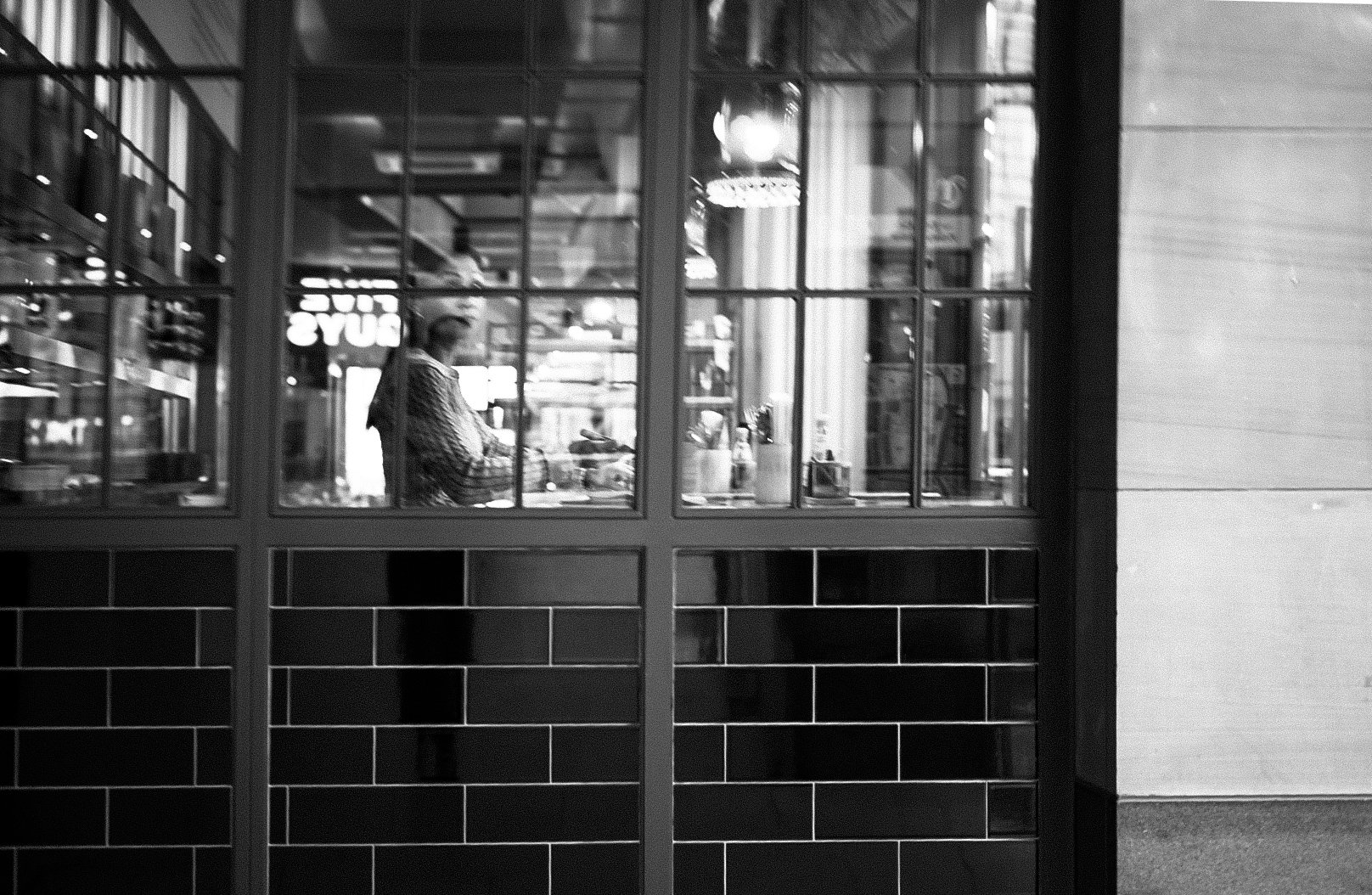 A black and white photo of a woman inside a coffee shop seen through a large window with grid panels. The woman is sitting at a counter, wearing a patterned shirt, and appears to be using a laptop or reading. Reflections of the street outside with bl