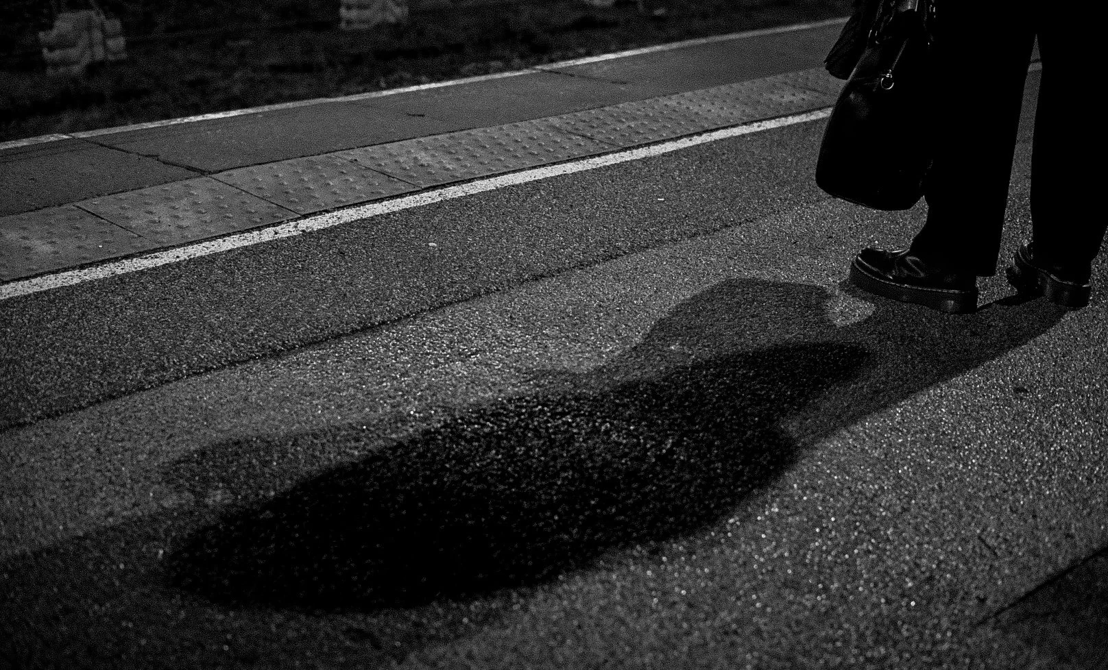Close-up of a person's legs and feet standing on a textured sidewalk at night, with their shadow cast on the ground.