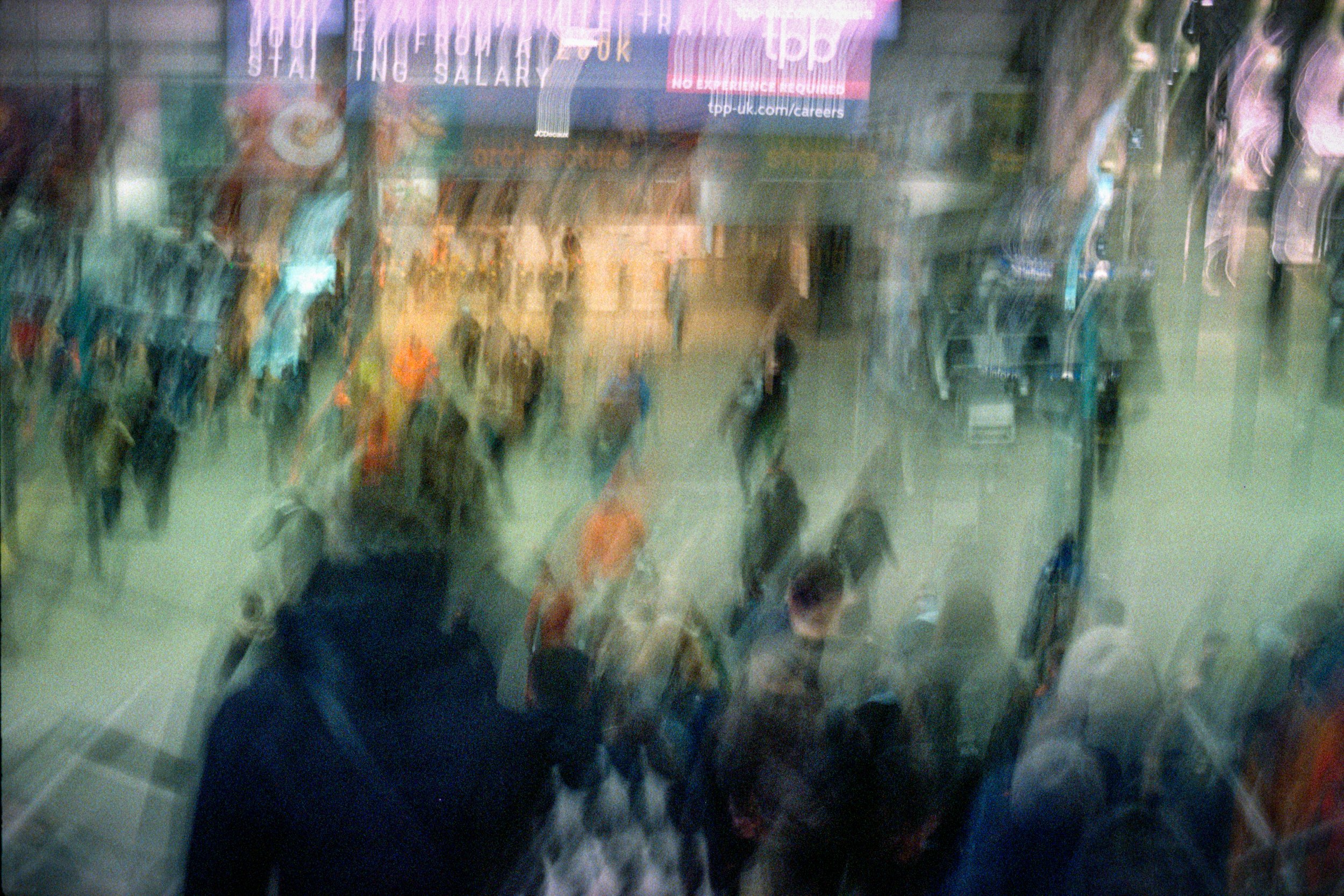 Blurred view of a busy street scene through a glass window, seen from inside a building, showing pedestrians walking outside.