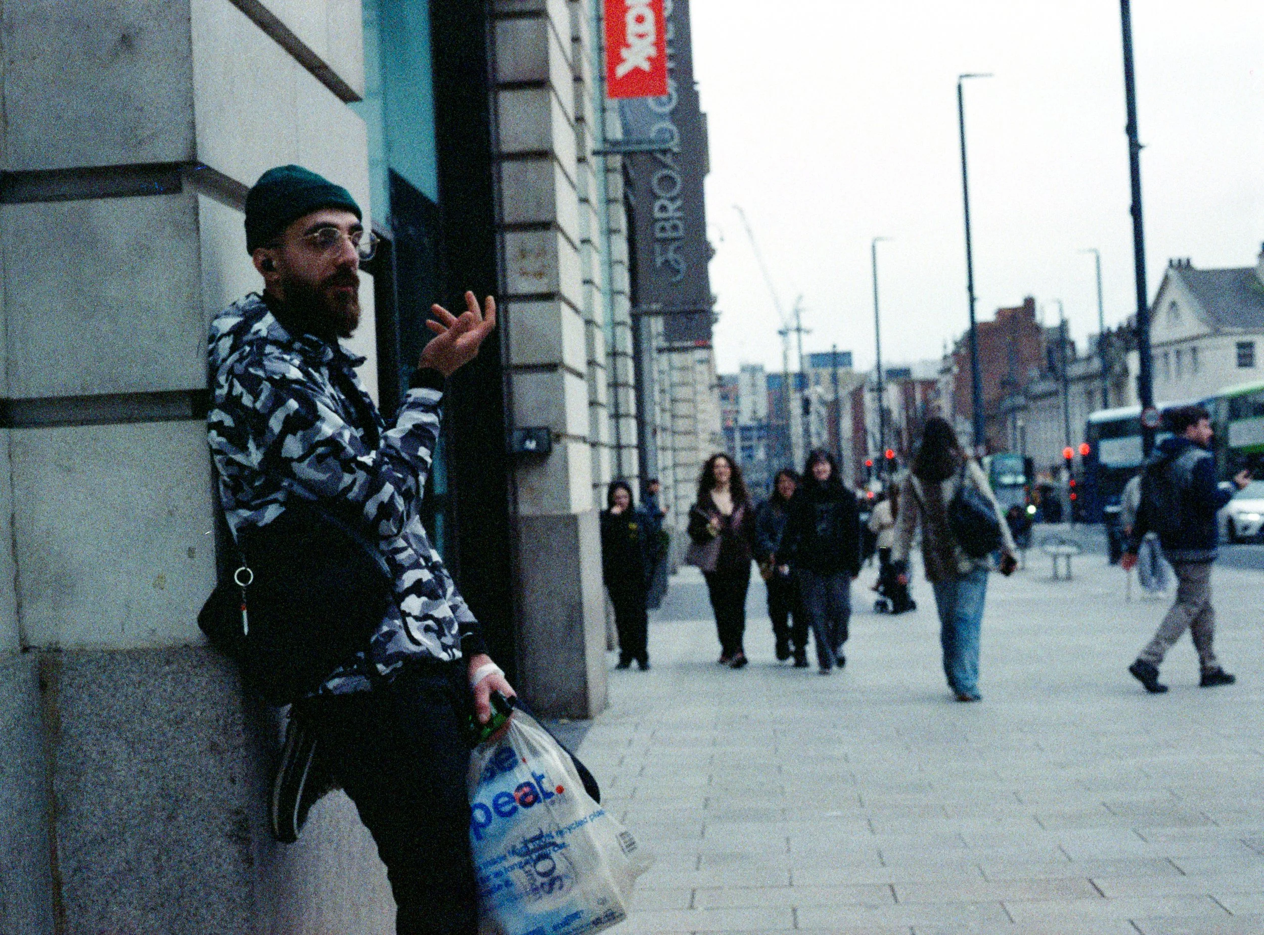A man with a dark beanie, glasses, and a patterned shirt pauses against a building while holding a plastic shopping bag on a busy city street with pedestrians and buses.
