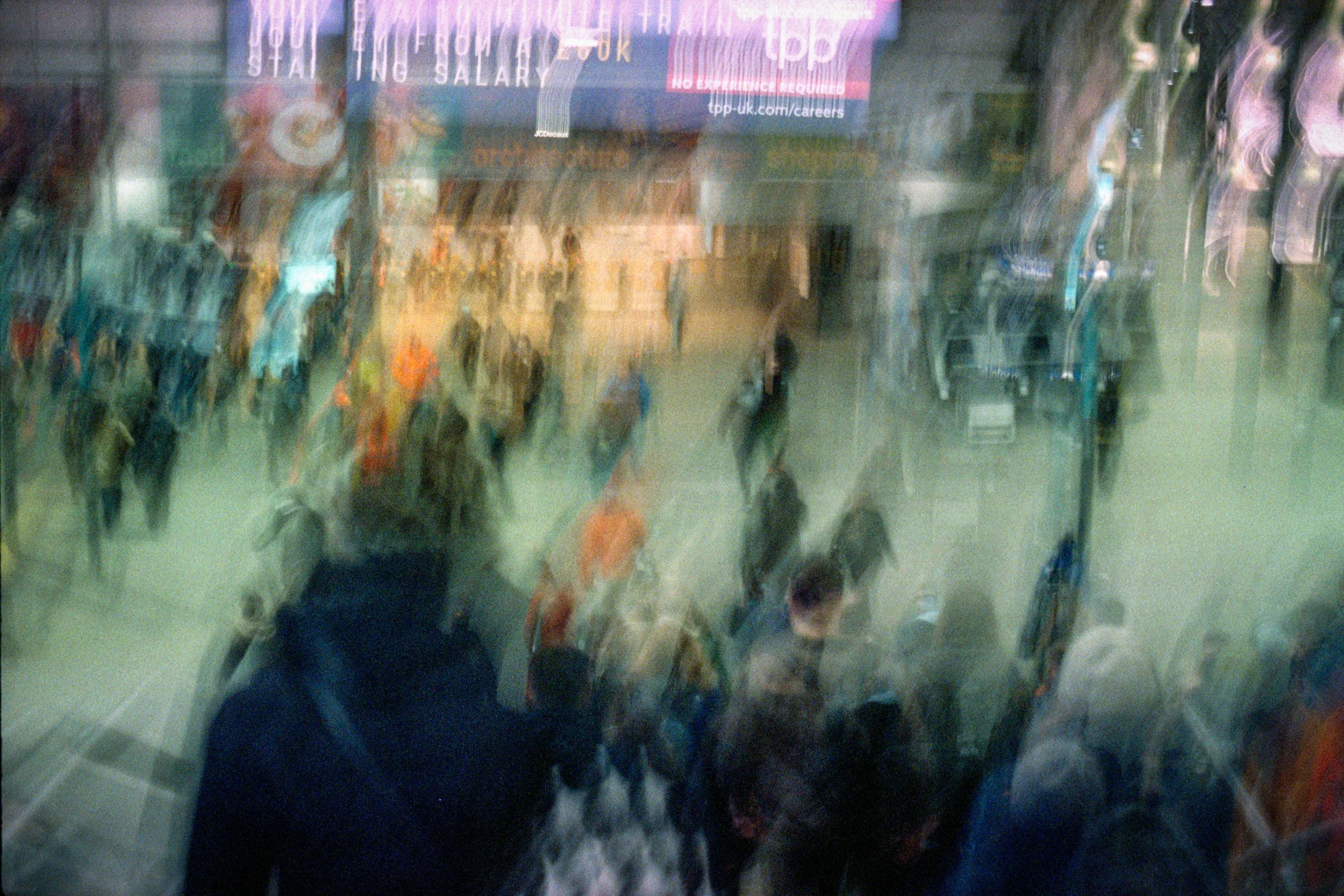 Blurred view of a busy city street with people walking, various shops, and illuminated signs at night.