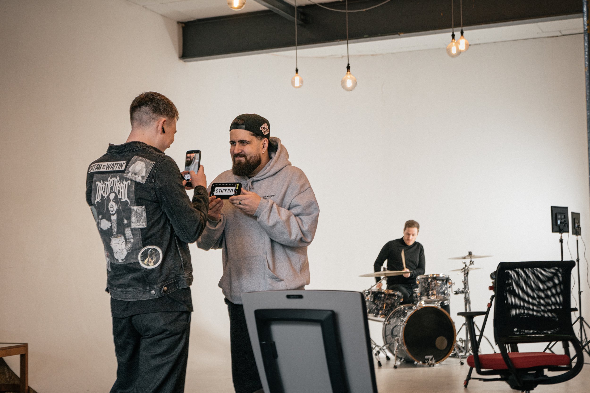 Two men are standing and taking a photo of each other with a smartphone inside a music studio or rehearsal space. One man is wearing a black jacket with patches and graphics, and the other man is wearing a gray hoodie and a black cap. In the backgrou