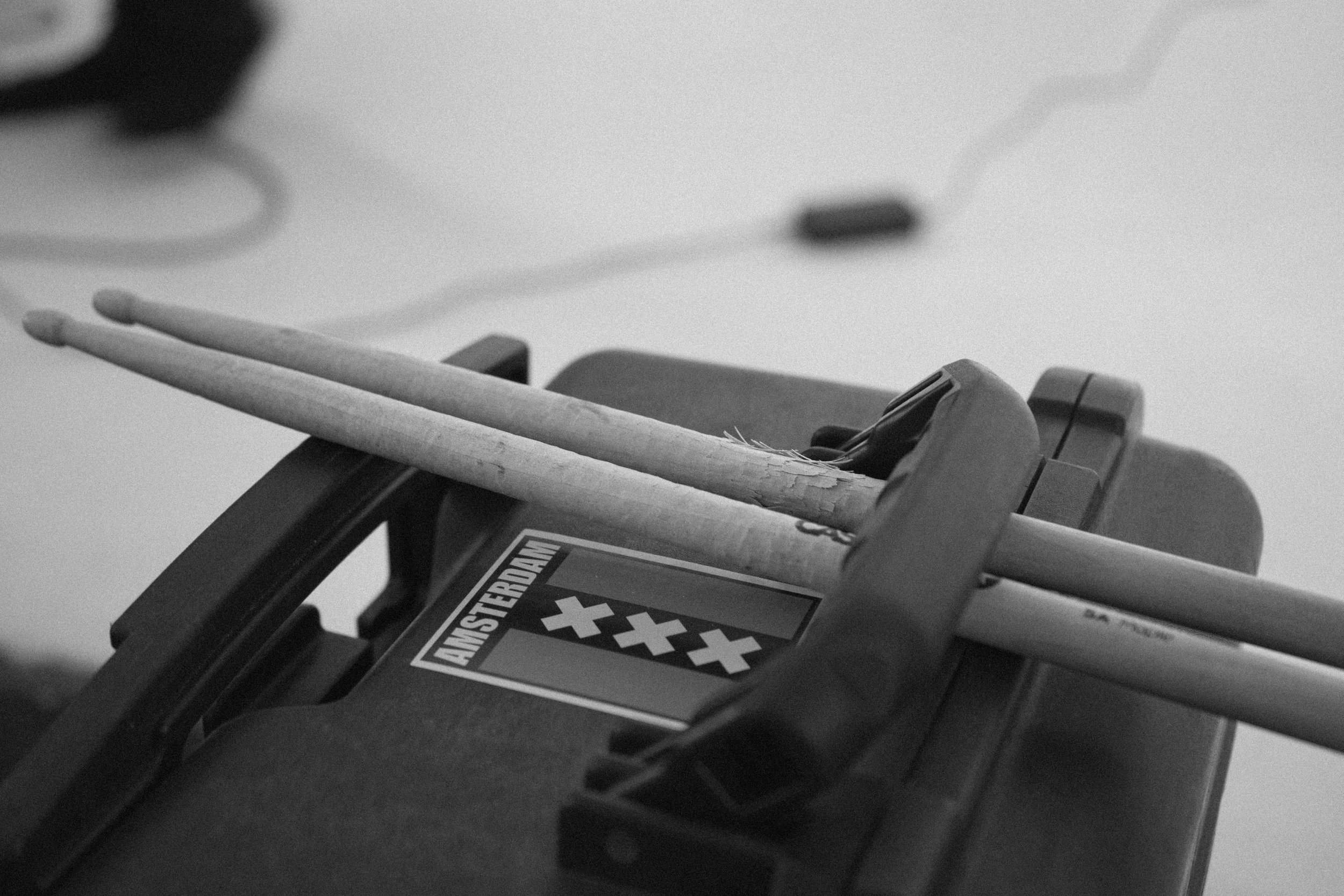 A pair of drumsticks resting on a black Amsterdam stickered object, with a black device and a blurred background.