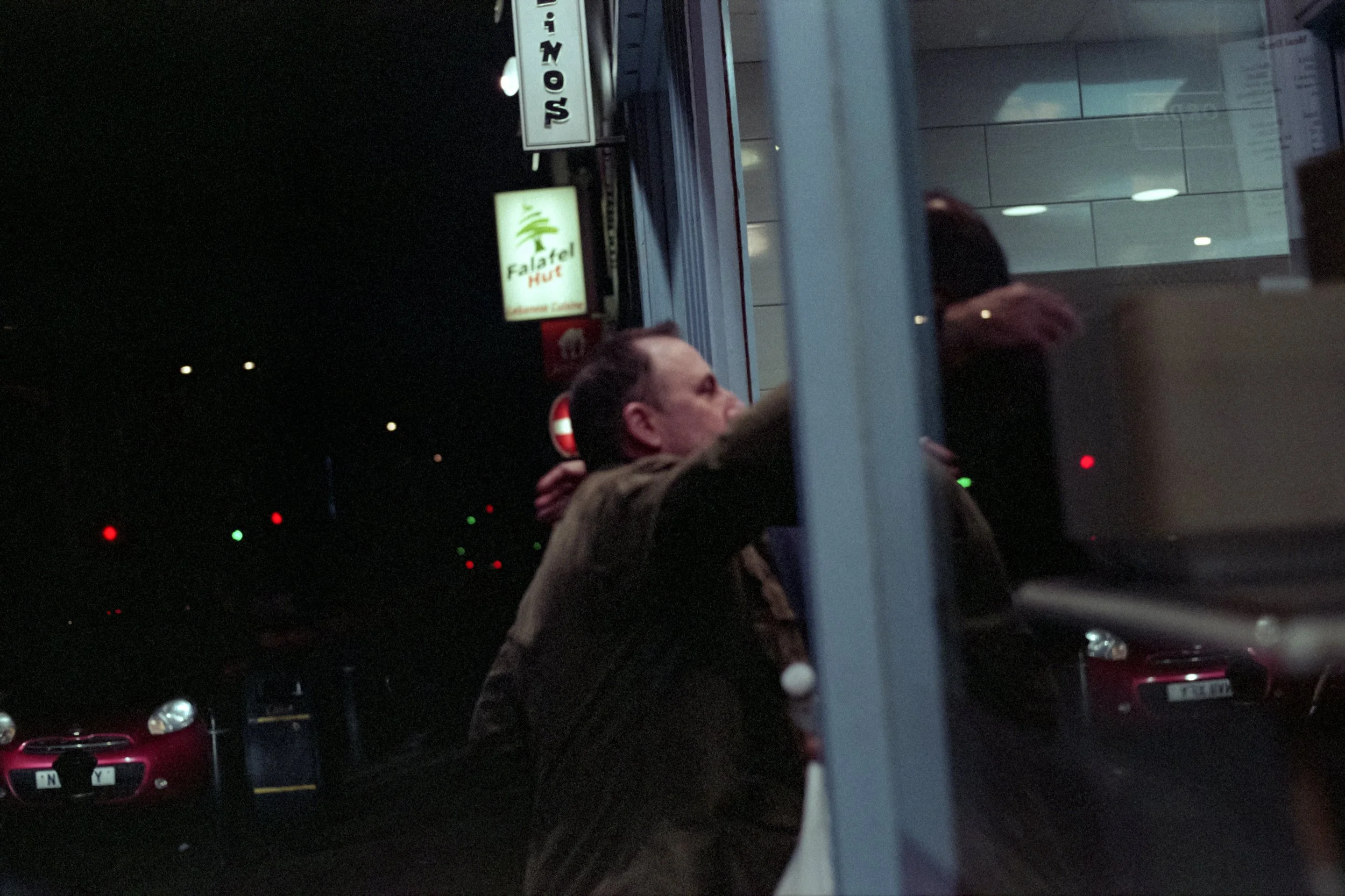 A man is standing outside at night, leaning against a glass window or door, with his arms behind his head. There are signs for a sushi restaurant and a falafel hut visible in the background, along with cars parked on the street.