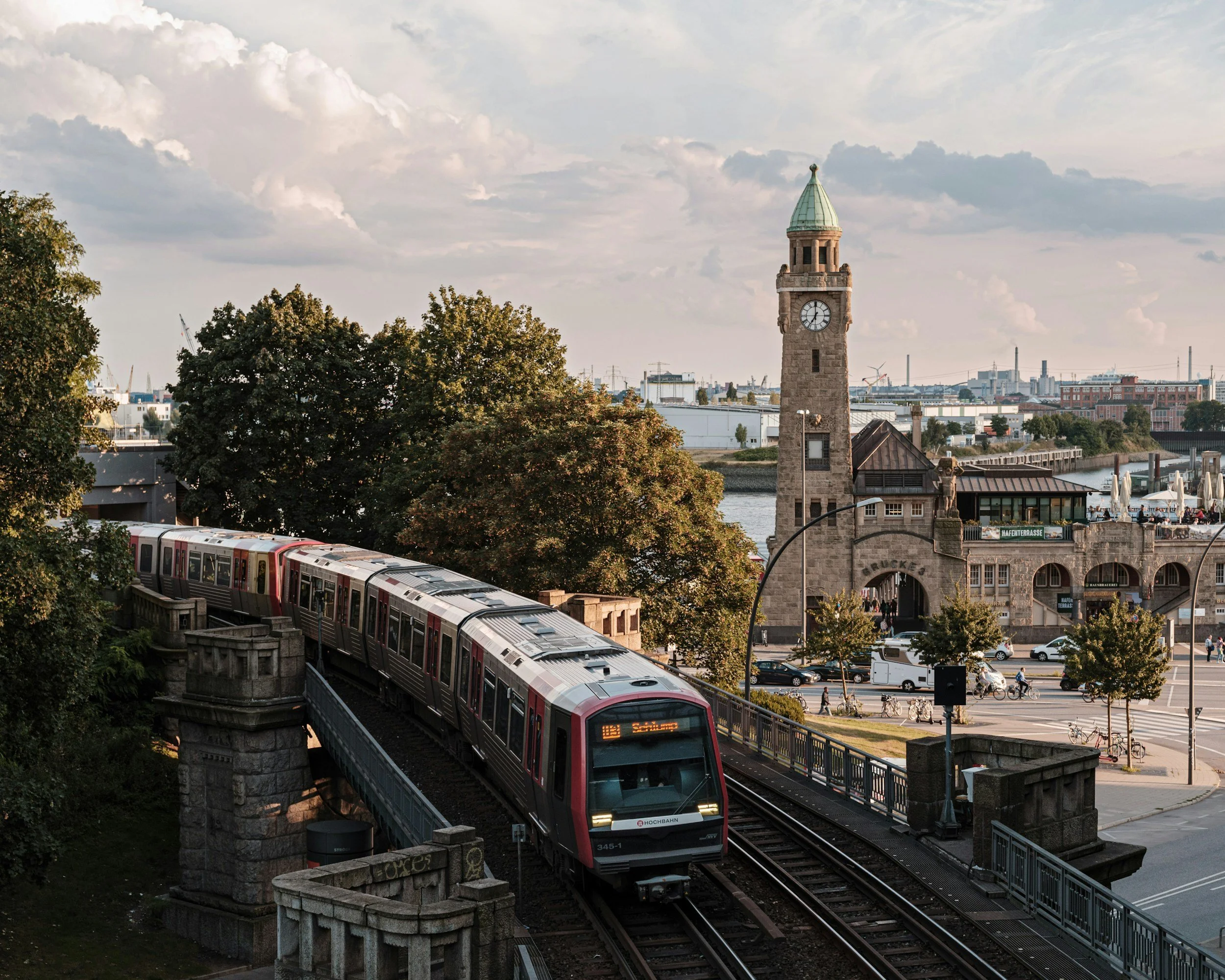 Un train rouge traverse un pont en pierre avec une tour d'horloge en arrière-plan, dans une ville au bord d'une rivière, avec des arbres et des bâtiments en arrière-plan.