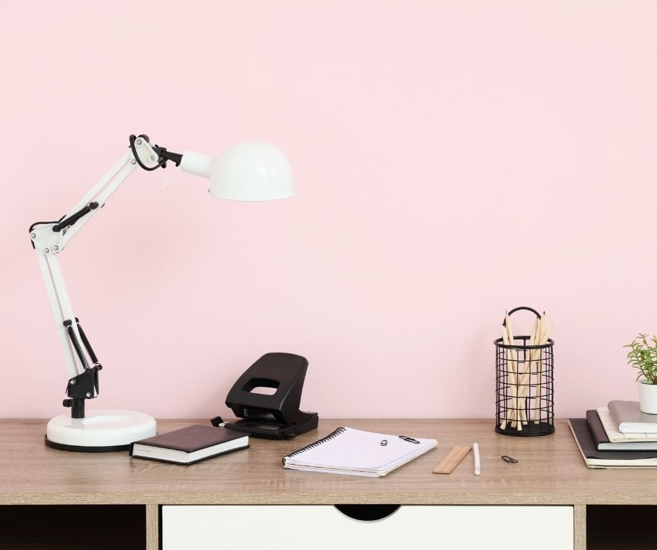 Bureau avec une lampe de bureau blanche, un massicot noir, un carnet noir, une pile de cahiers, un cahier blanc avec un stylo noir, une règle en bois, un pinceau, un trombone, un pot à crayons en métal noir avec des crayons, un pot avec une plante en