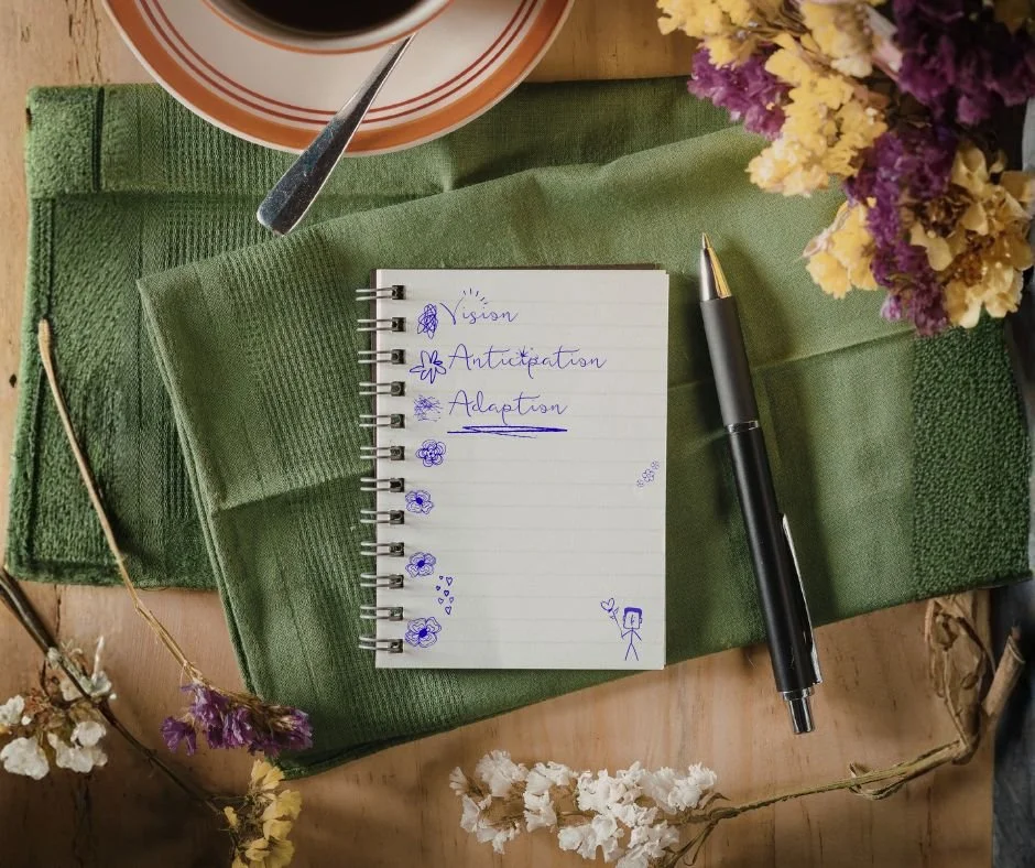 Une nappe verte, un carnet à spirale avec des mots écrits en français, un stylo noir, des fleurs séchées violettes et blanches, une tasse de café en haut, une cuillère, et une assiette avec une boisson.