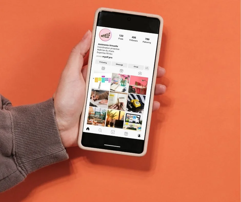 Main tenant un smartphone affichant le profil Instagram d'une account nommée MYELF, avec une mise en page de photo de profil rose, noms et statistiques. La personne porte un pull gris clair et la photo est prise sur un fond orange.