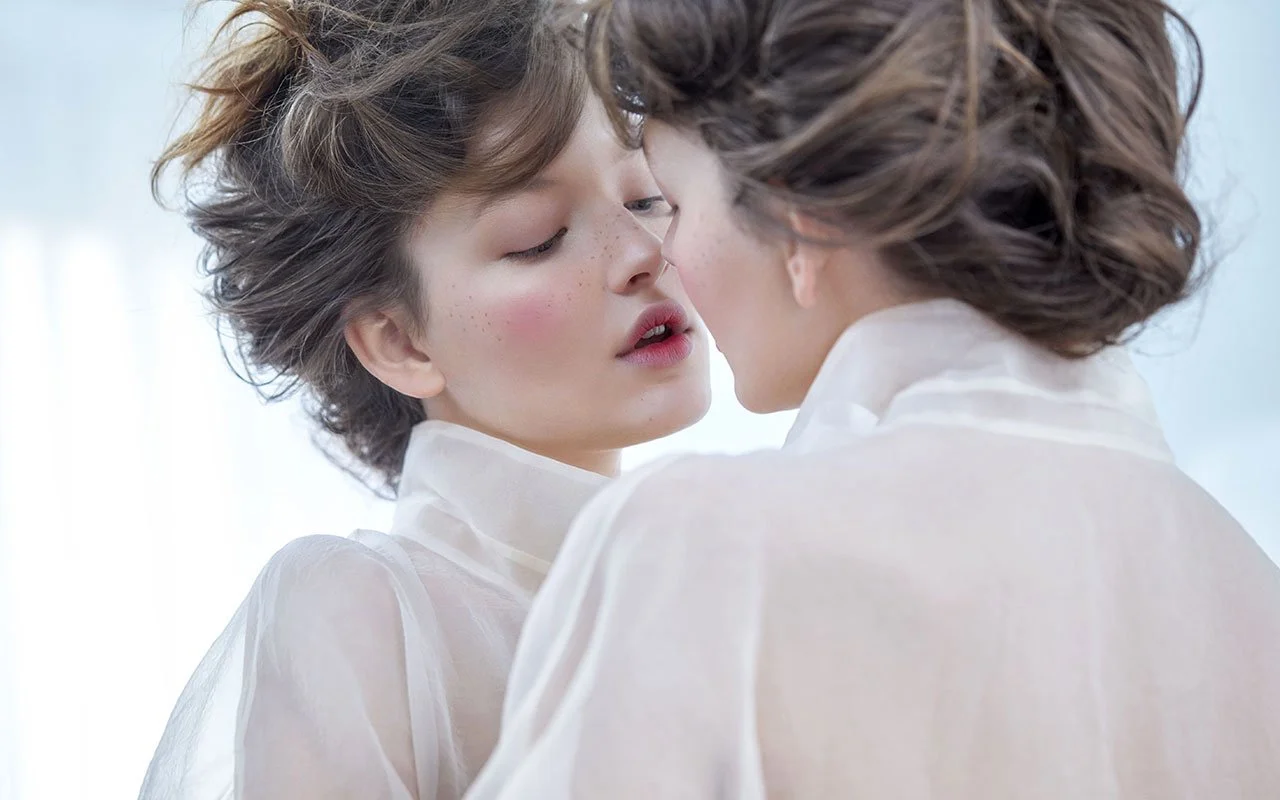Deux femmes aux cheveux bouclés, se rapprochant presque pour s'embrasser, portant des vêtements légers et translucides dans un environnement lumineux.