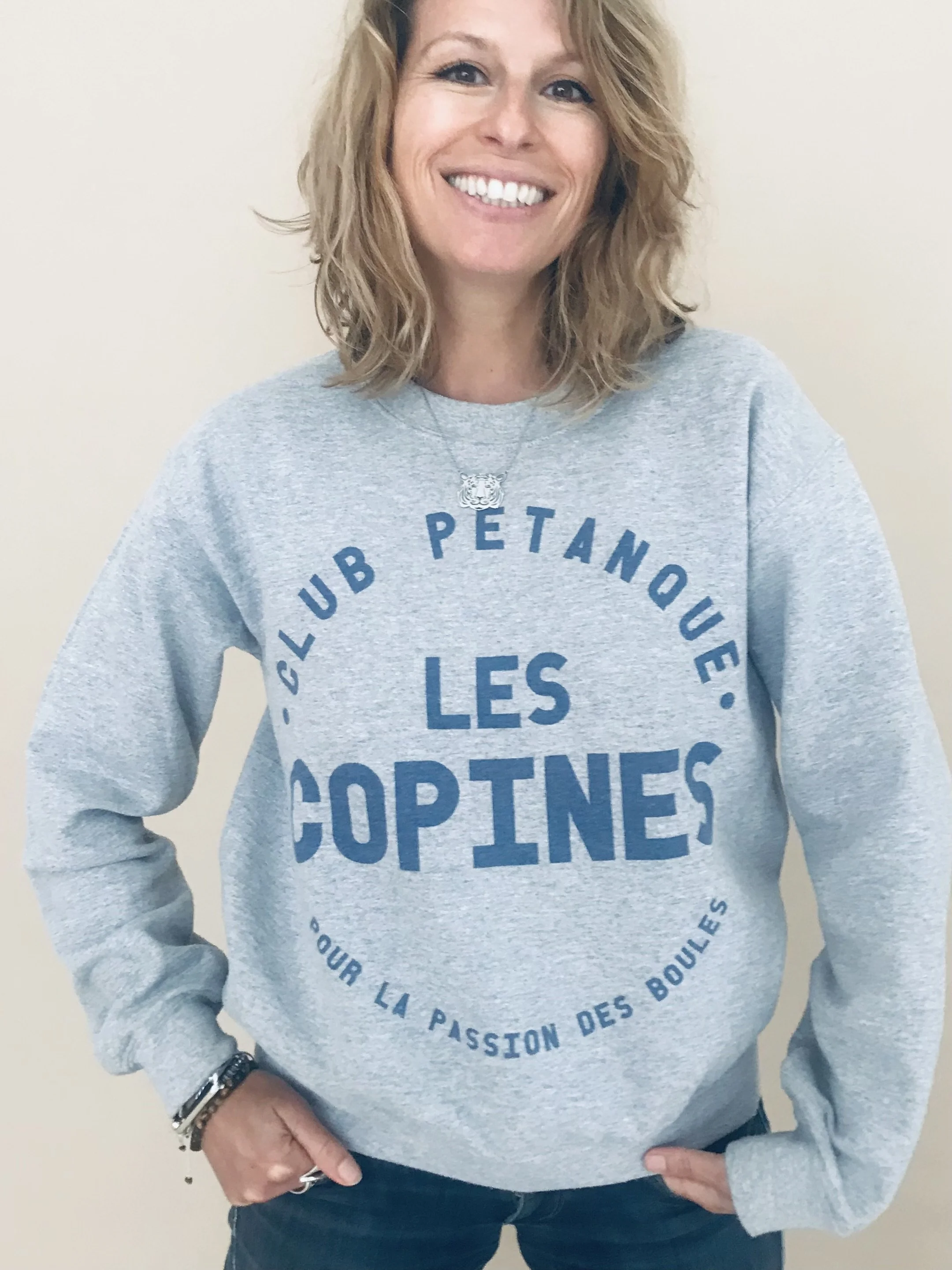 Femme souriante portant un sweat gris avec le texte en bleu 'Club Pétanque Les Copines', se tenant contre un mur neutre.