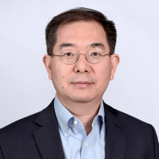 A man with glasses wearing a dark suit and light blue shirt, posing against a plain light background.
