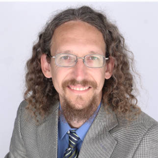 Portrait of a man with long curly hair and glasses, wearing a gray suit and blue shirt with a patterned tie, smiling against a light background.