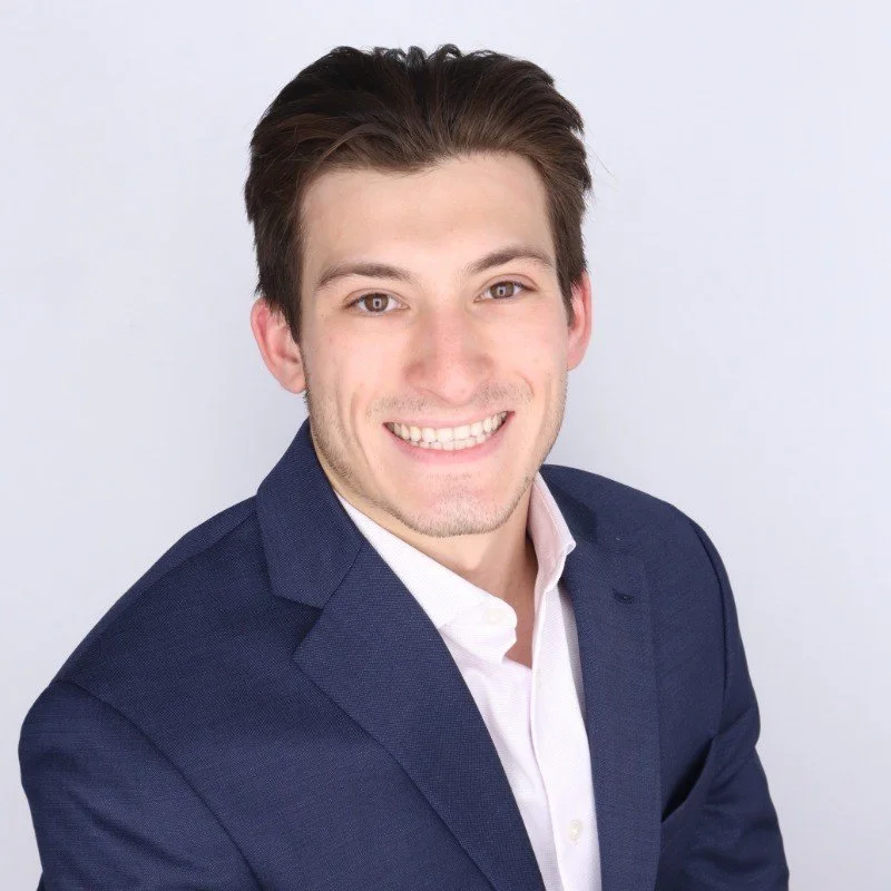 Portrait of a young man with dark hair, wearing a navy blazer and white shirt, smiling against a light background.