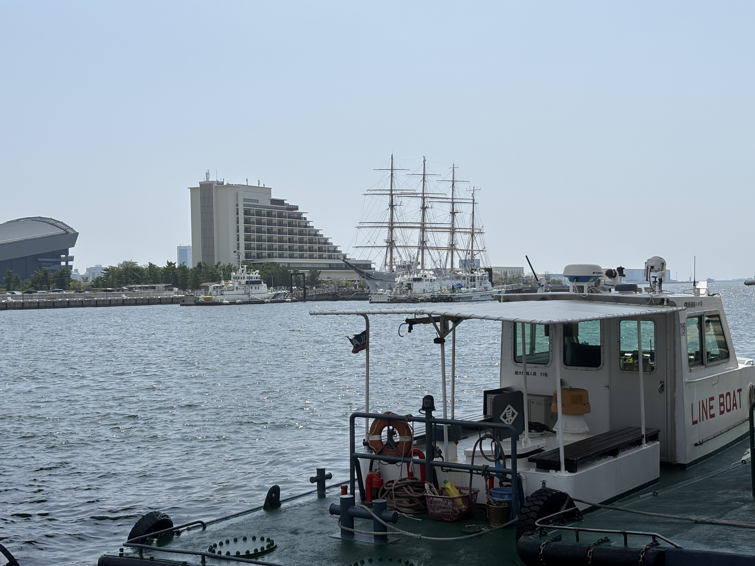 Ein Hafen mit mehreren Segelschiffen und einem Motorboot im Vordergrund, moderne Gebäude am Ufer, ruhiges Wasser, blauer Himmel.