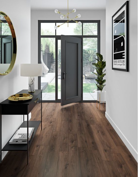 Modern entryway with open gray door, large window, and hardwood flooring. Decor includes a black console table with a white lamp and gold tray, a potted plant in a white stand, a round gold wall mirror, and a framed black-and-white poster on the white wall.