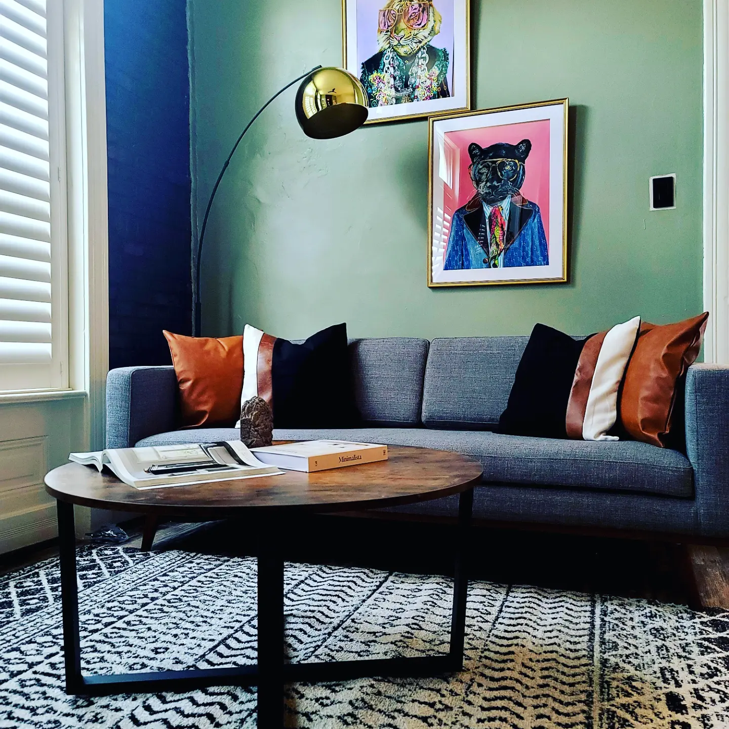 Living room with a gray sofa accented by four pillows in black, white, and brown, a wooden coffee table with books and a decorative stone, a patterned rug, green wall with framed colorful animal portraits, a large floor lamp with a gold shade, and a window with white shutters.