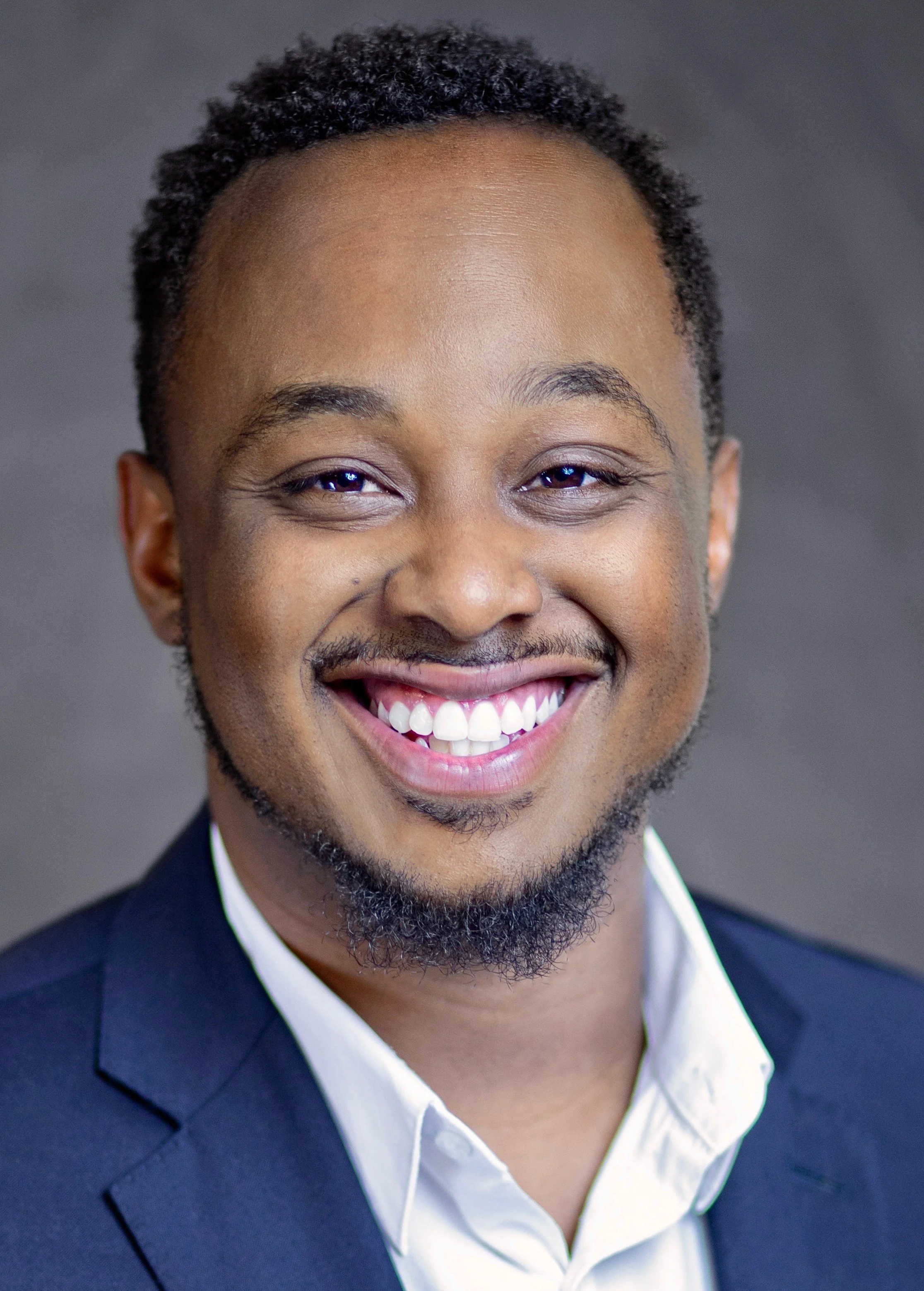 Close-up of a smiling man in a suit and white shirt against a grey background.