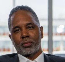 Close-up of a middle-aged man with short, curly hair and a beard, wearing a dark suit and tie, sitting indoors near a window.