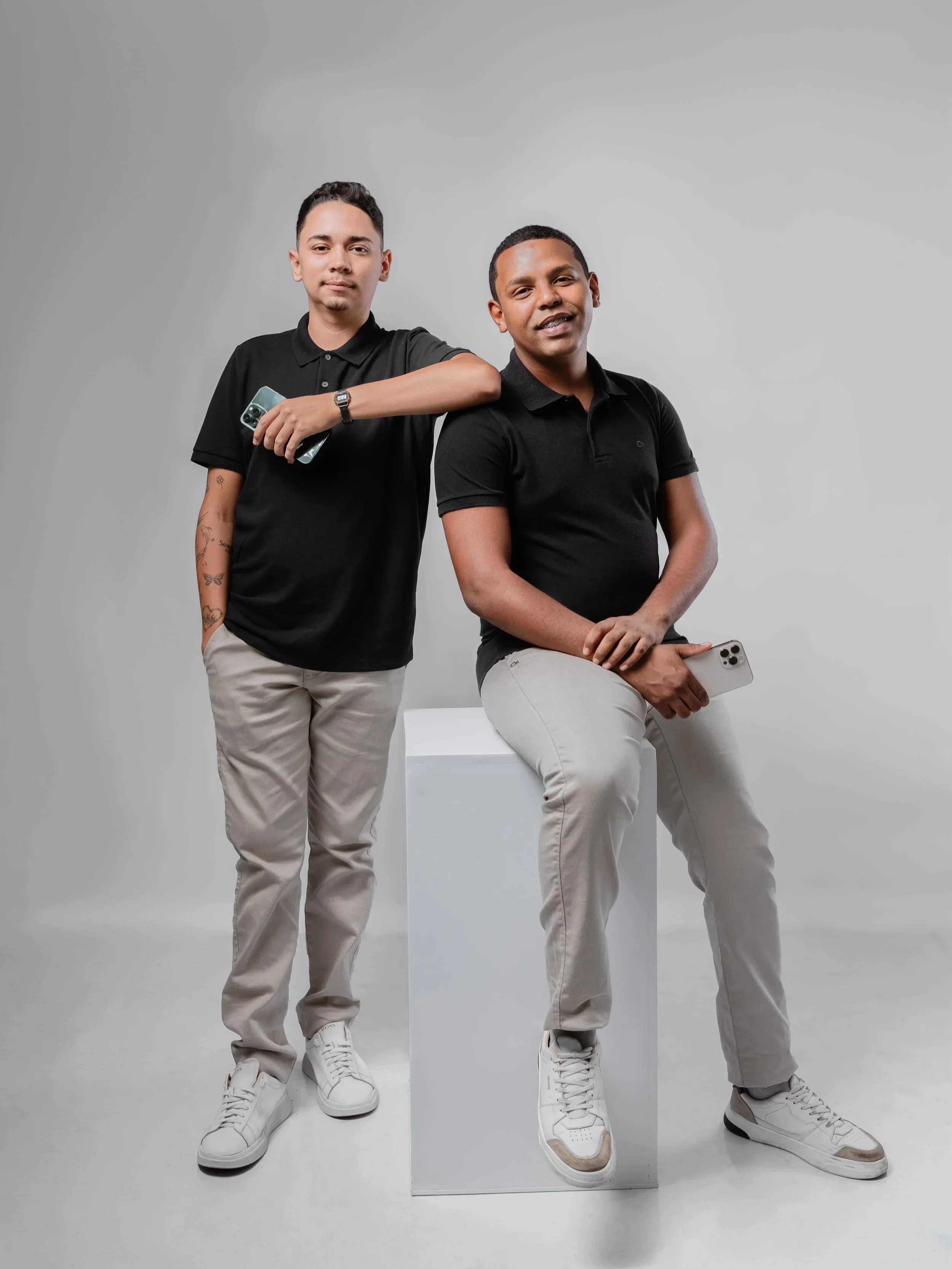 Dois homens jovens com roupas pretas e calças bege, um segurando um celular e o outro sentado em um bloco branco, ambos segurando celulares, em um fundo cinza.