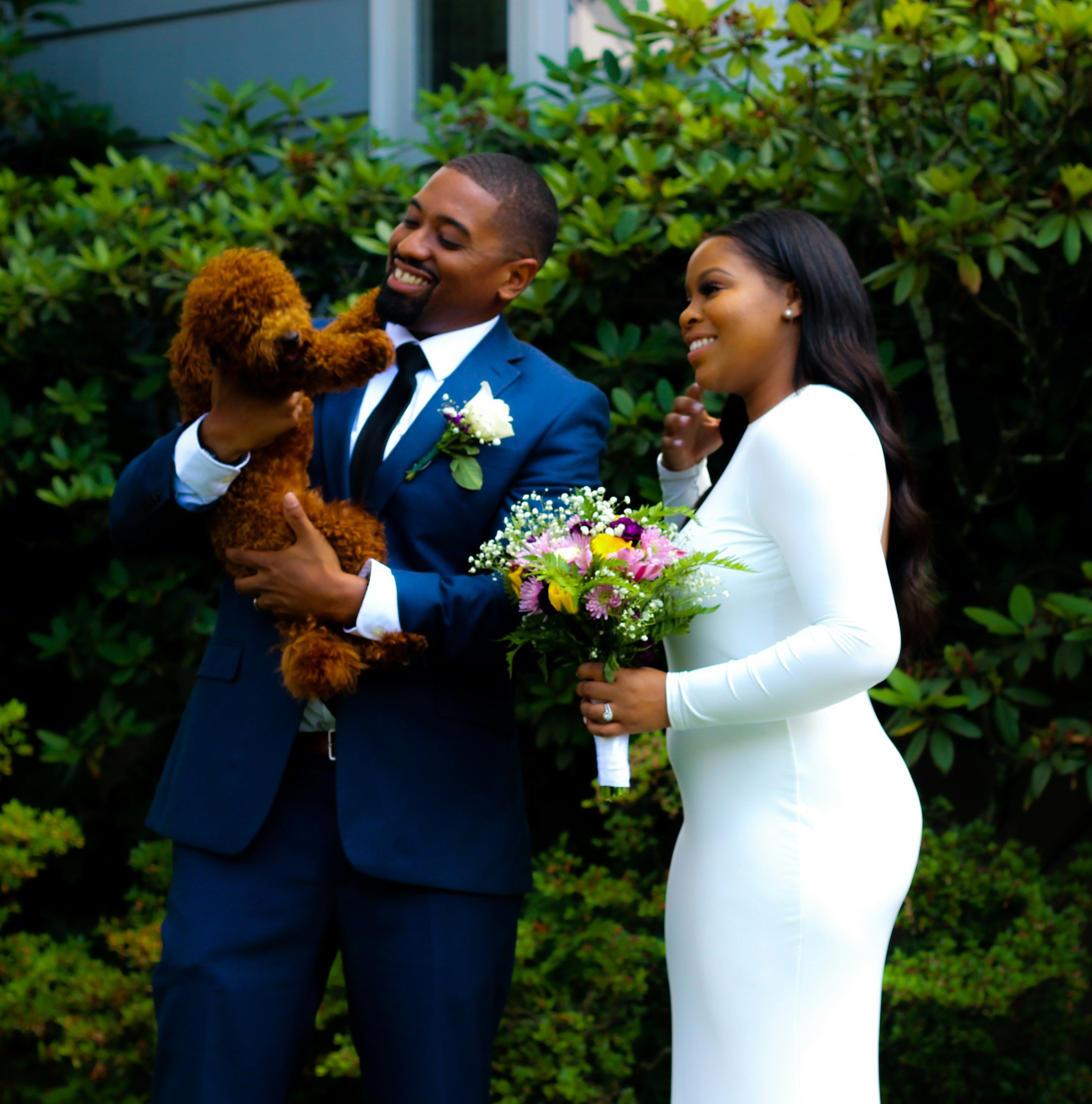 A couple in wedding attire standing outdoors, with the groom holding a small brown poodle and the bride holding a colorful bouquet, surrounded by green bushes.