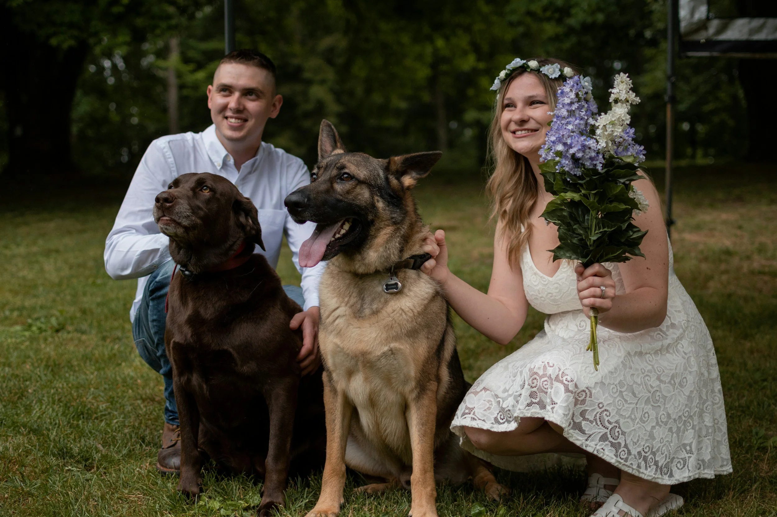 A couple with two dogs outdoors, woman holding a flowers bouquet, man kneeling behind a chocolate lab, woman with a blonde braid and white dress with floral crown, two dogs sitting on grass, large trees in background.
