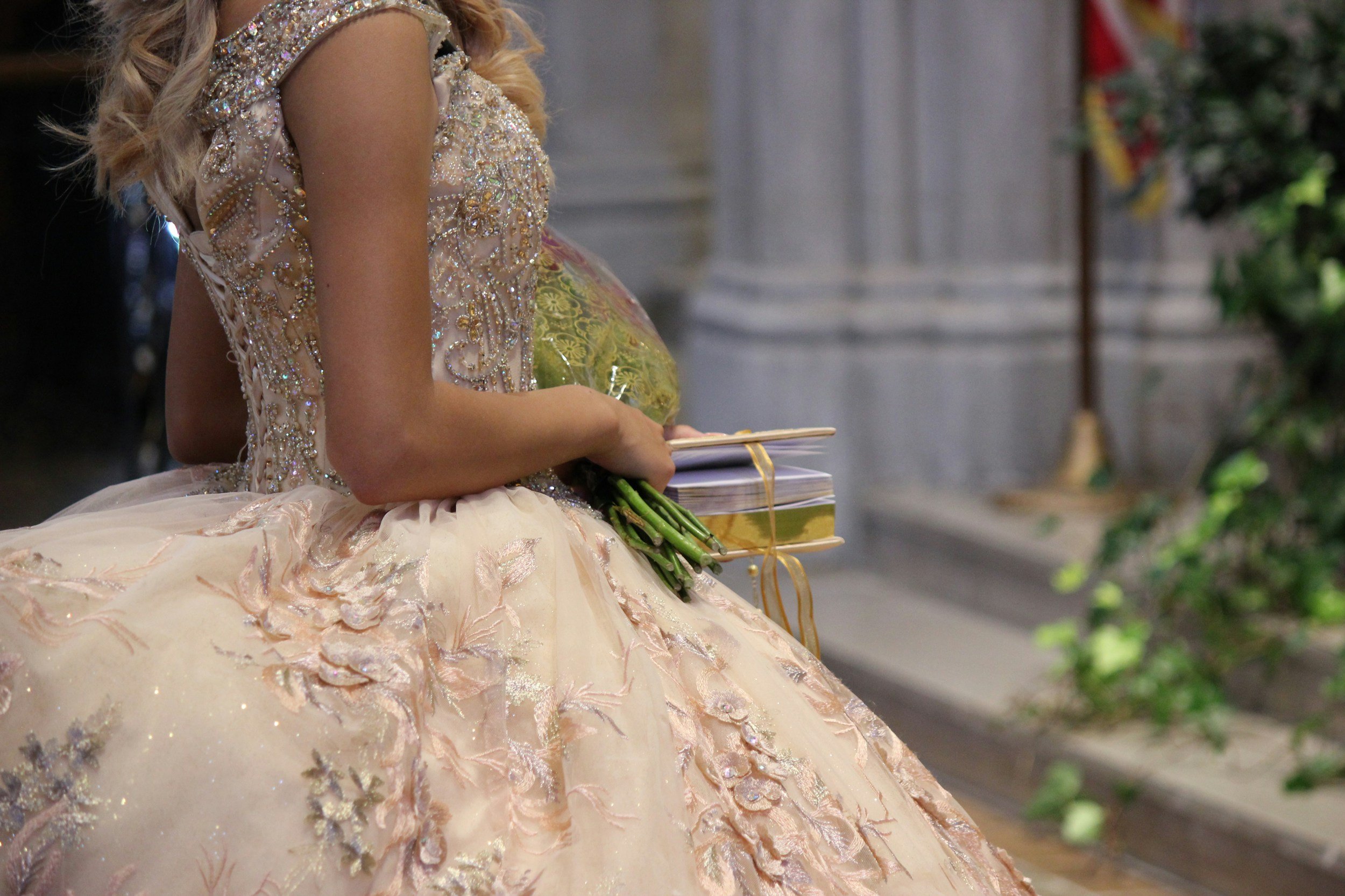 Part of a woman in an ornate, beaded dress holding a small bouquet of green flowers and two sealed packages tied with a golden ribbon. She is sitting on a bench in a formal setting with marble columns and greenery.