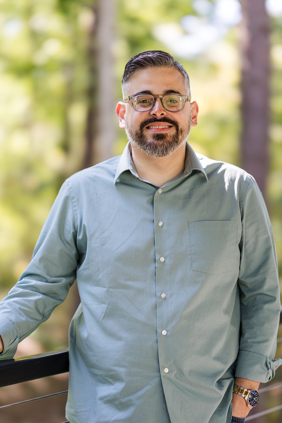 A man with glasses and a beard standing outdoors on a sunny day, wearing a light green button-up shirt and a watch, with trees and green foliage in the background.