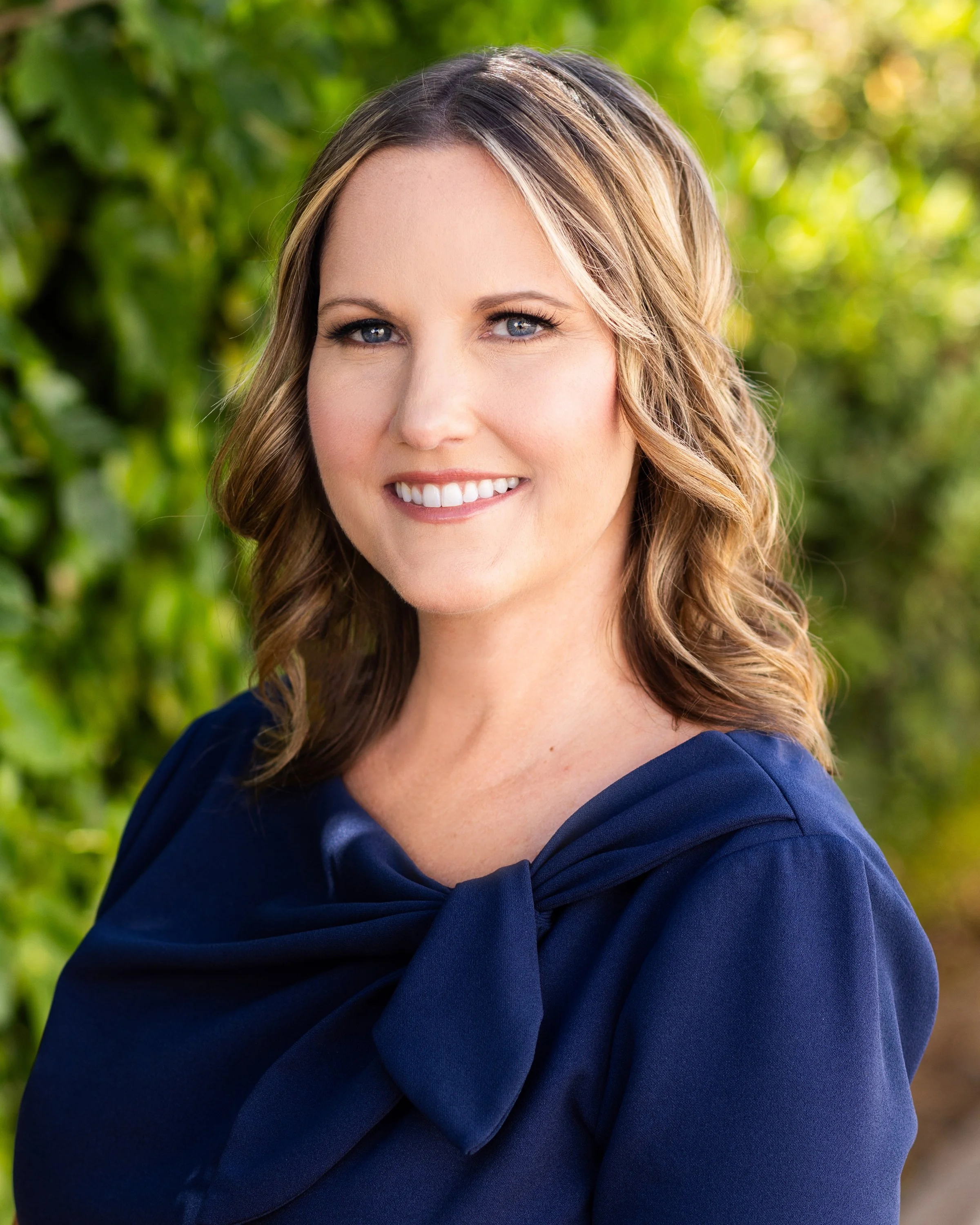A smiling woman with shoulder-length wavy brown hair, wearing a navy blue top with a bow detail on the shoulder, standing outdoors with green foliage in the background.