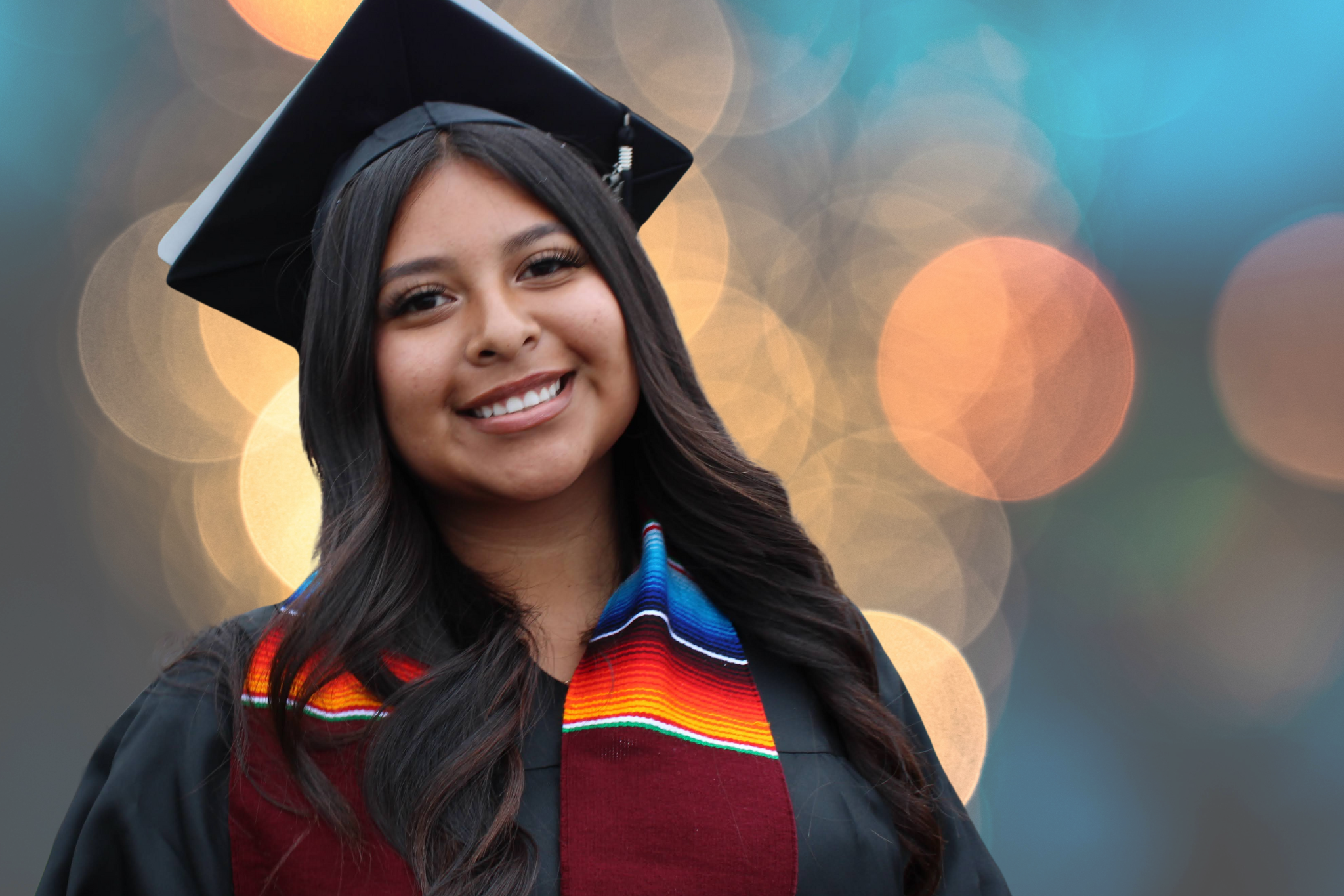 A young woman in a graduation gown and cap, smiling, with a colorful stole around her neck, standing against a blurred background of lights.