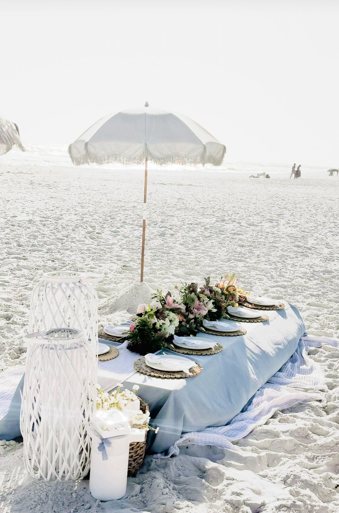 Beach setup with a table decorated with flowers, plates, and glasses, under a white beach umbrella, on sandy shore with few people and umbrellas in the background.