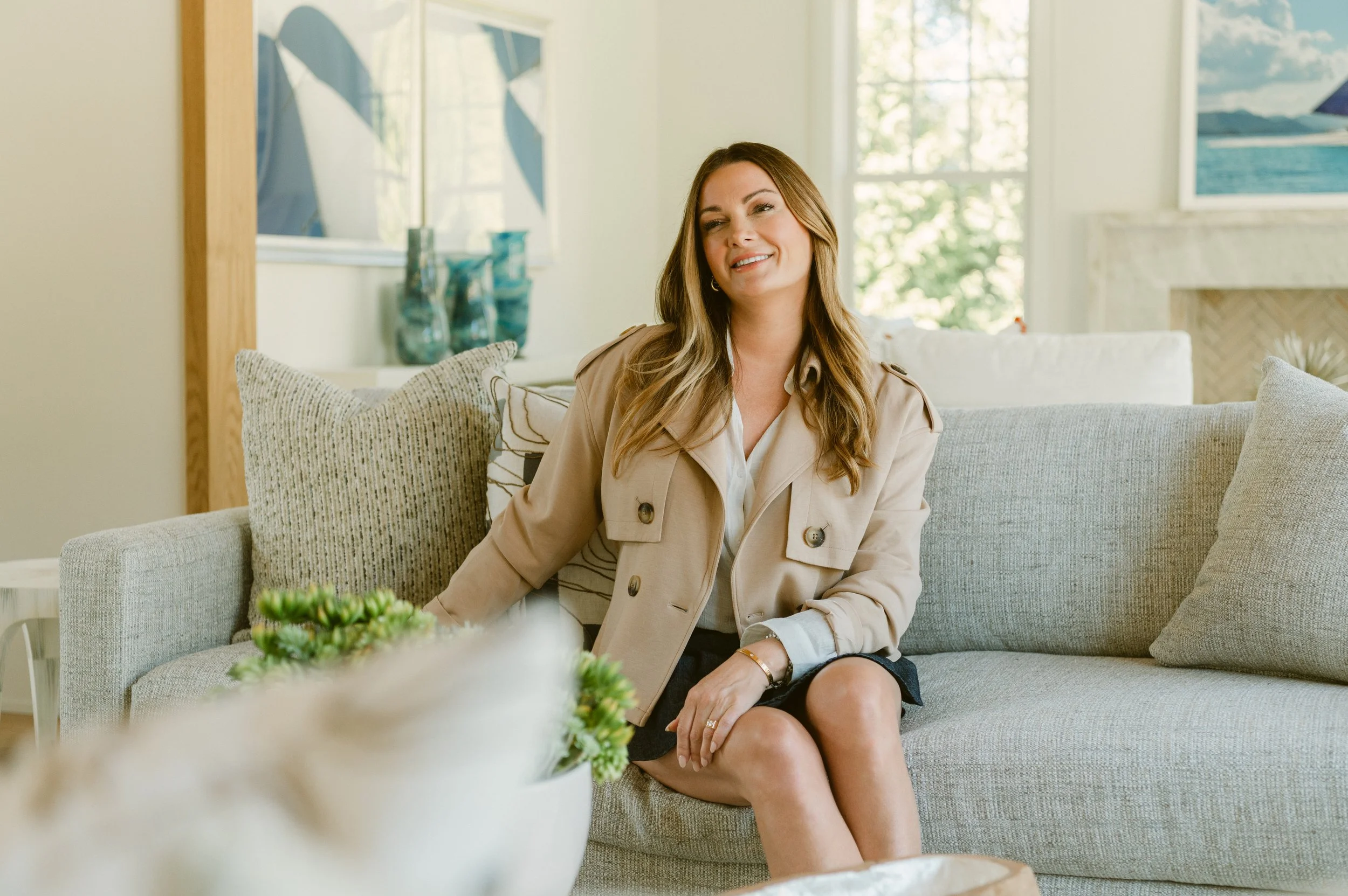Lisa sitting on a light-colored couch in a bright living room, smiling and looking to her left. She has long, wavy brown hair and is wearing a beige trench coat and a black skirt.